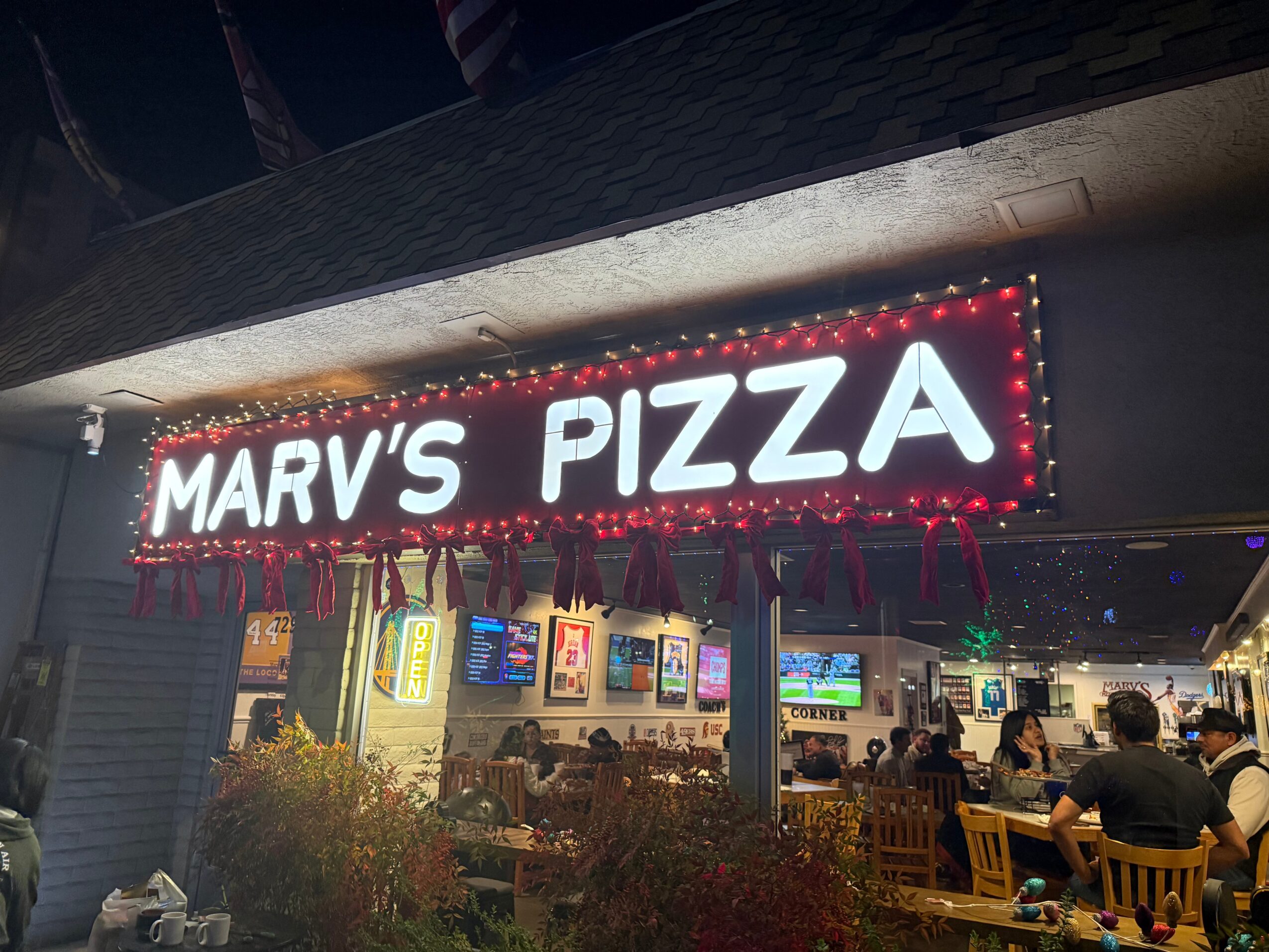 Bright sign reads 'MARV'S PIZZA' above a restaurant interior with people dining and multiple screens.