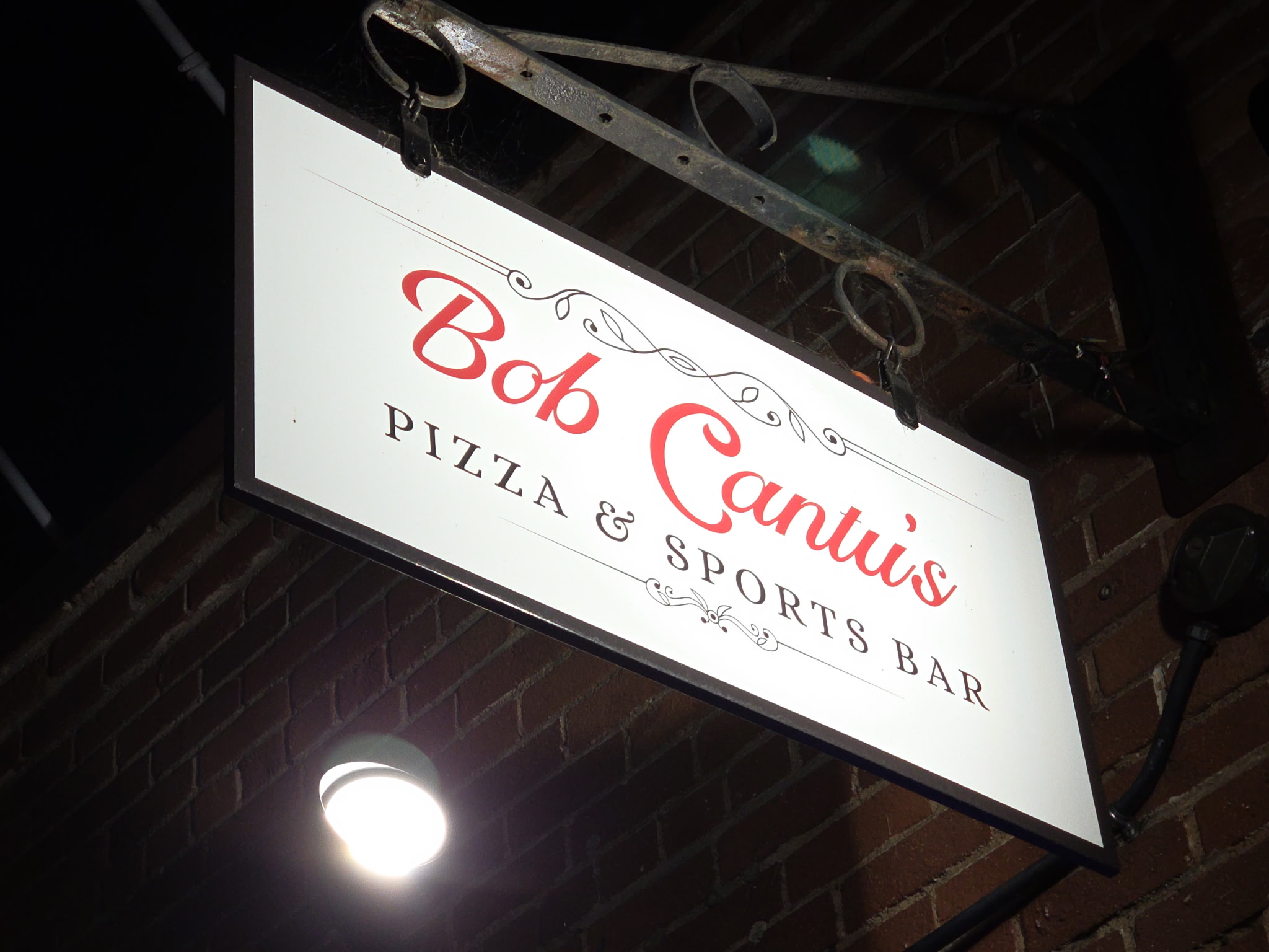 Sign for Bob Canuis Pizza & Sports Bar hanging from ceiling, with brick wall background.