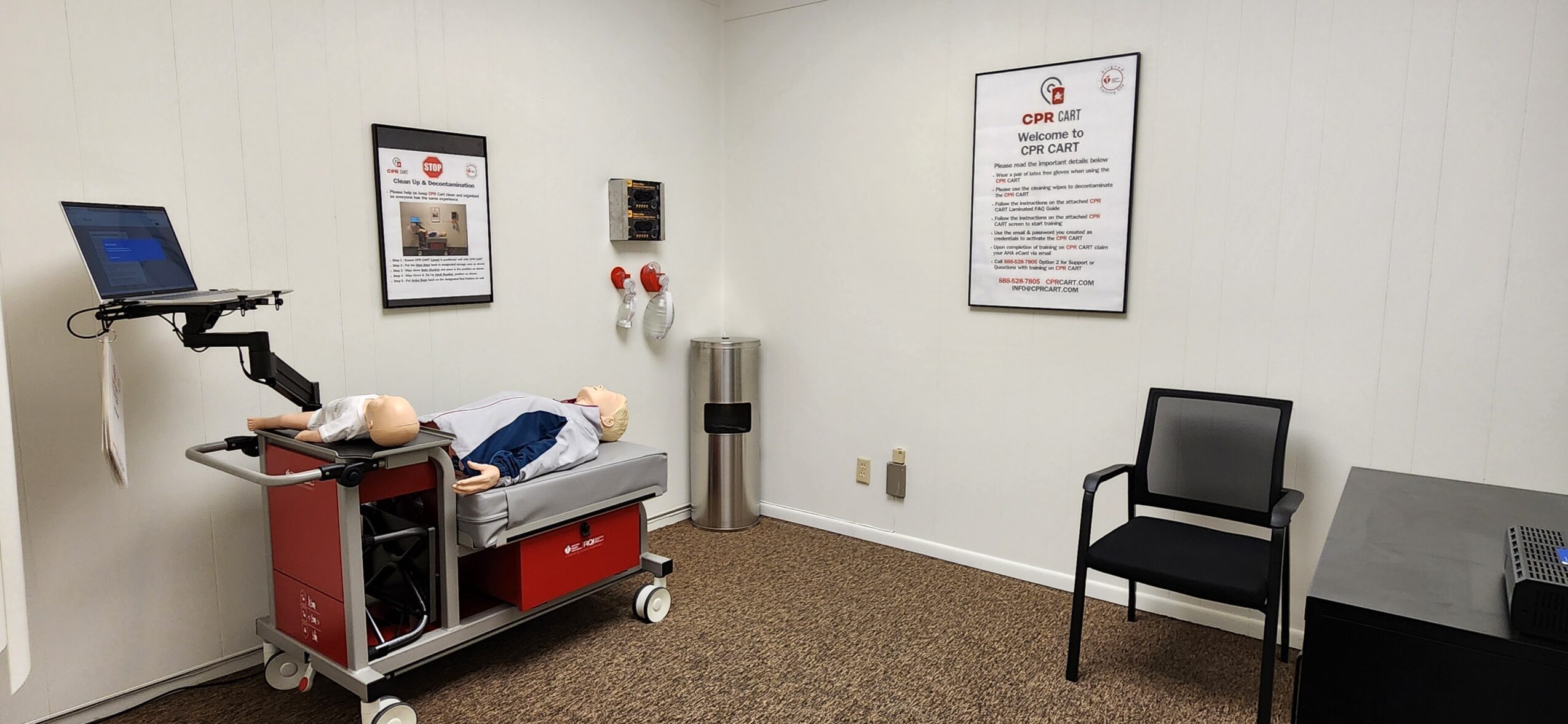 Hospital room with a bed, medical equipment, a chair, and wall posters, with white walls and brown carpet.