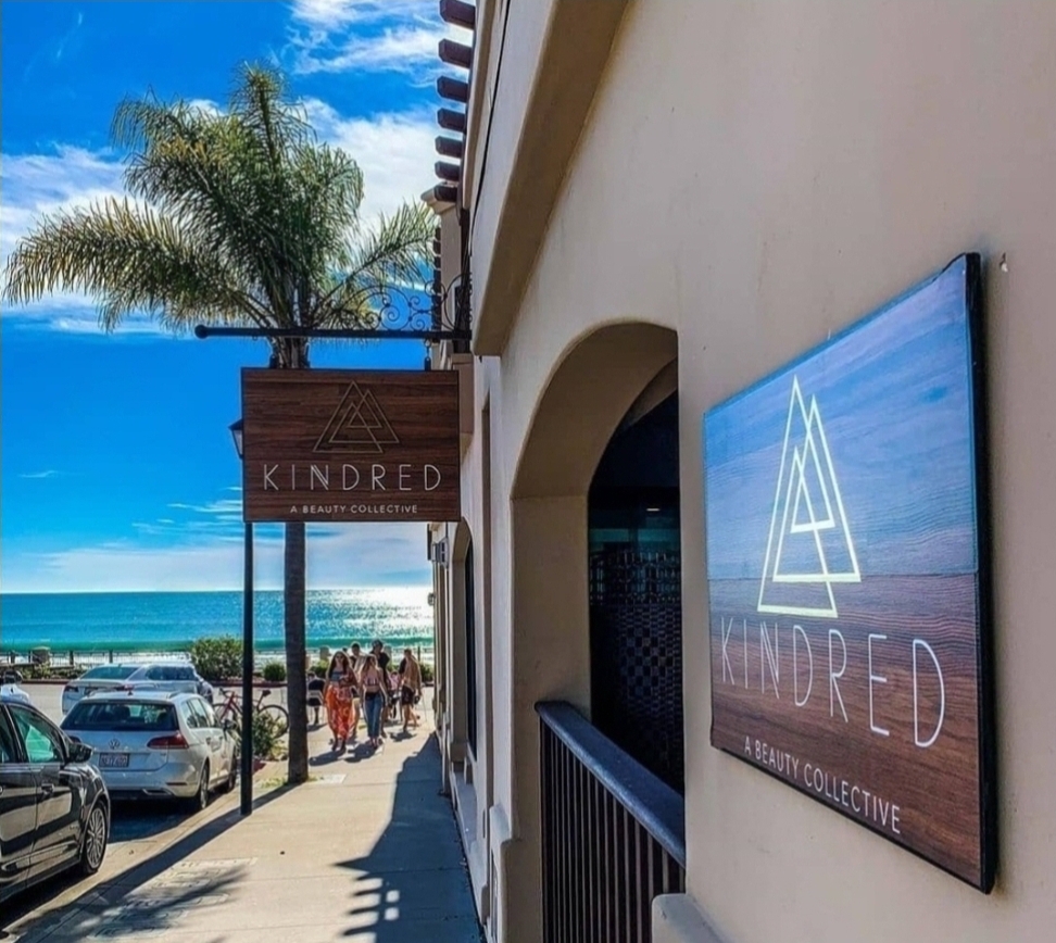 Street view with palm tree, parked cars, and a sign for Kindred, a health and wellness collective, near a beach.