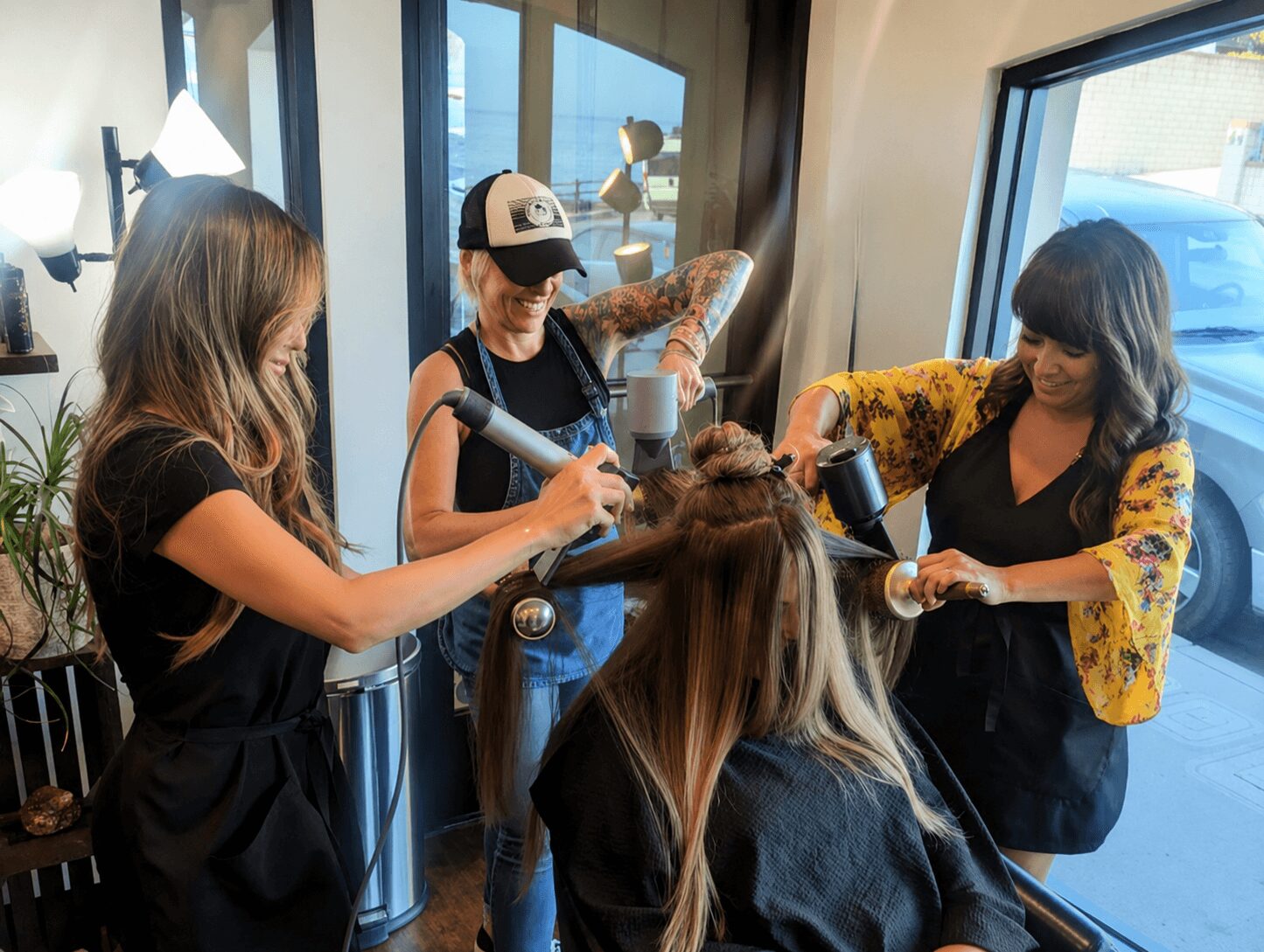 Three women in a hair salon, one styling a client's hair, another with a hairdryer, and a third observing, near a large window.