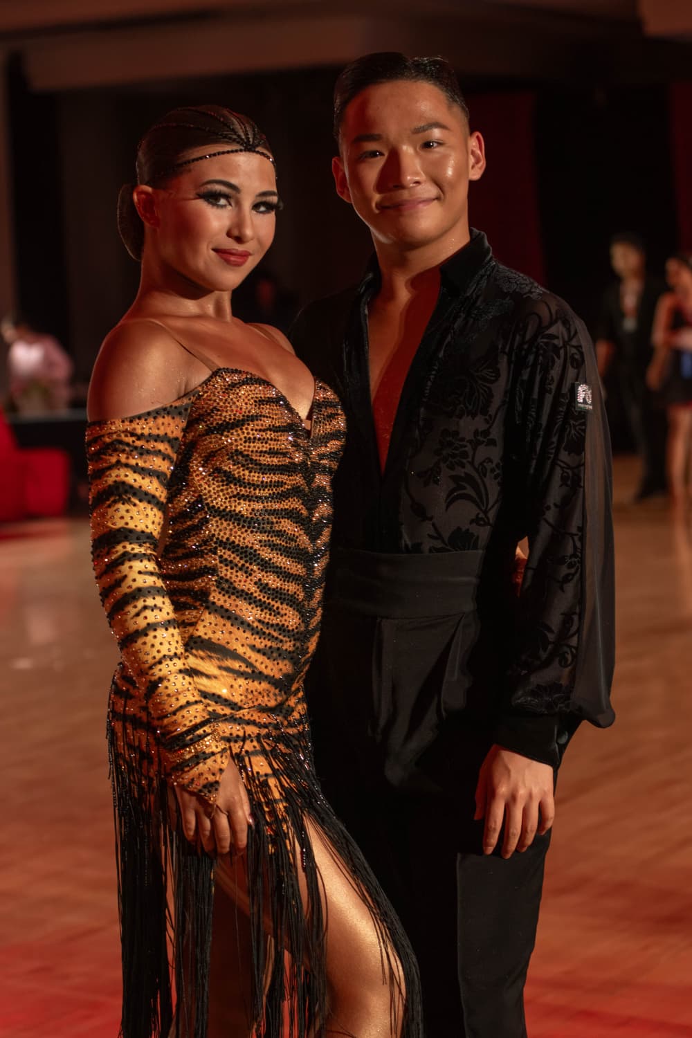 A woman and a man pose together in a dance setting, dressed in elegant dance attire, with a blurred background.