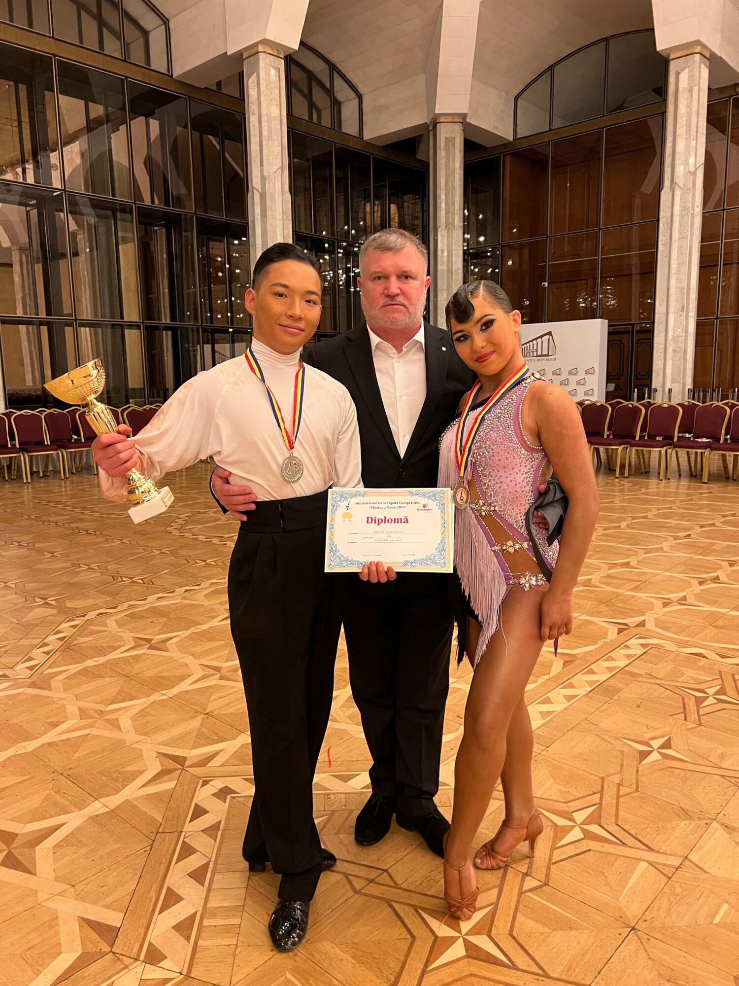 Three people standing indoors, one holding a certificate and flowers, with a wooden floor and glass windows in the background.