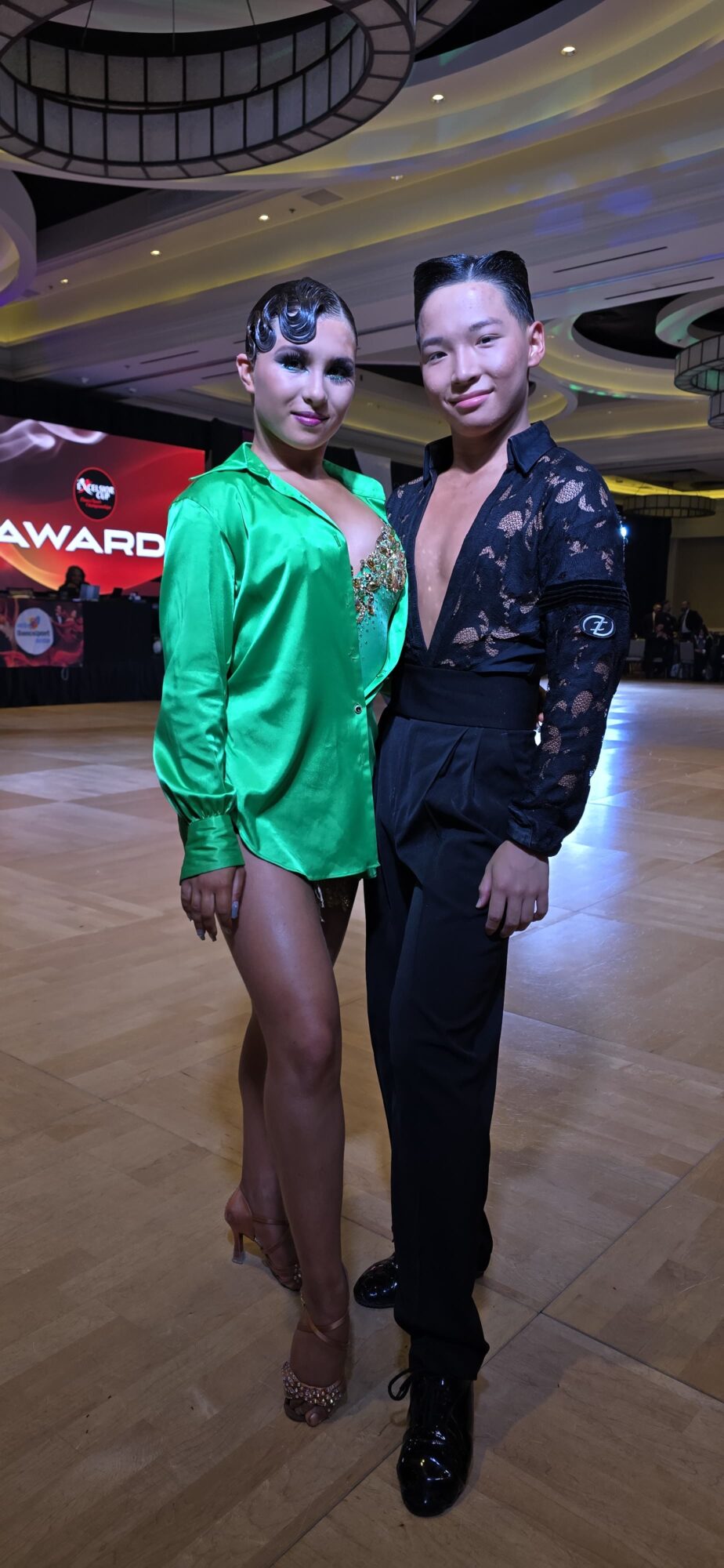 Two women pose together in a ballroom with a wooden floor and a large screen in the background.