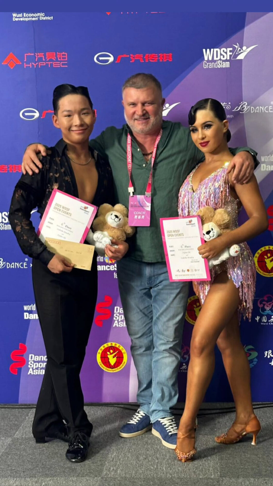 Three people standing together holding certificates and teddy bears, posing in front of a blue backdrop with logos.