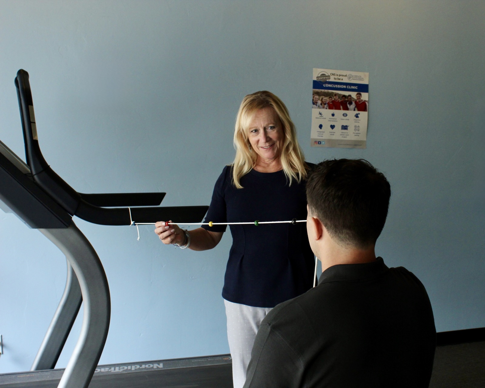 Woman holding a measuring tape in front of a man, standing near a treadmill, against a blue wall with a poster.