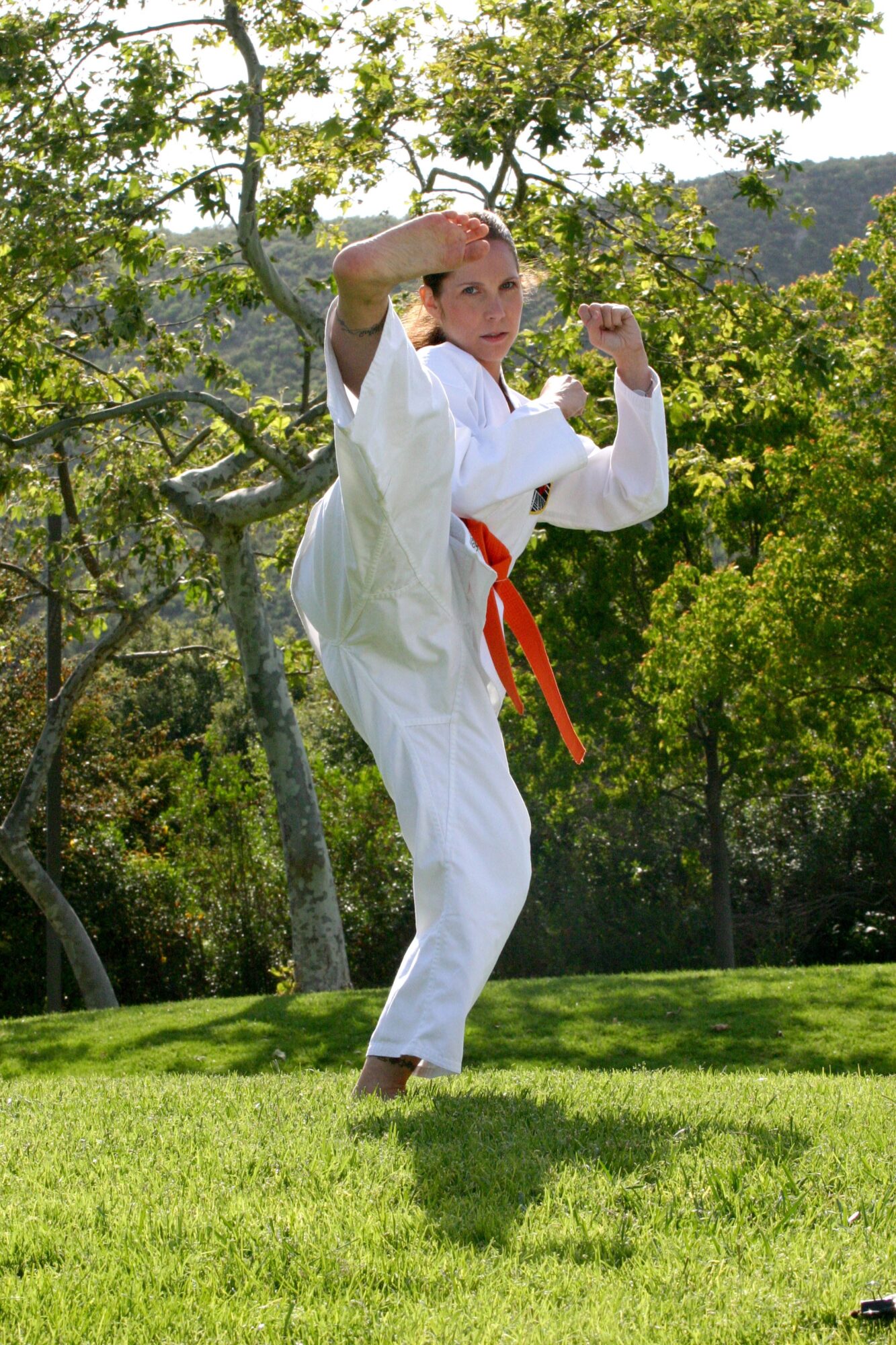 Young person in a white martial arts uniform with an orange belt practicing a stance outdoors on grass, trees in background.