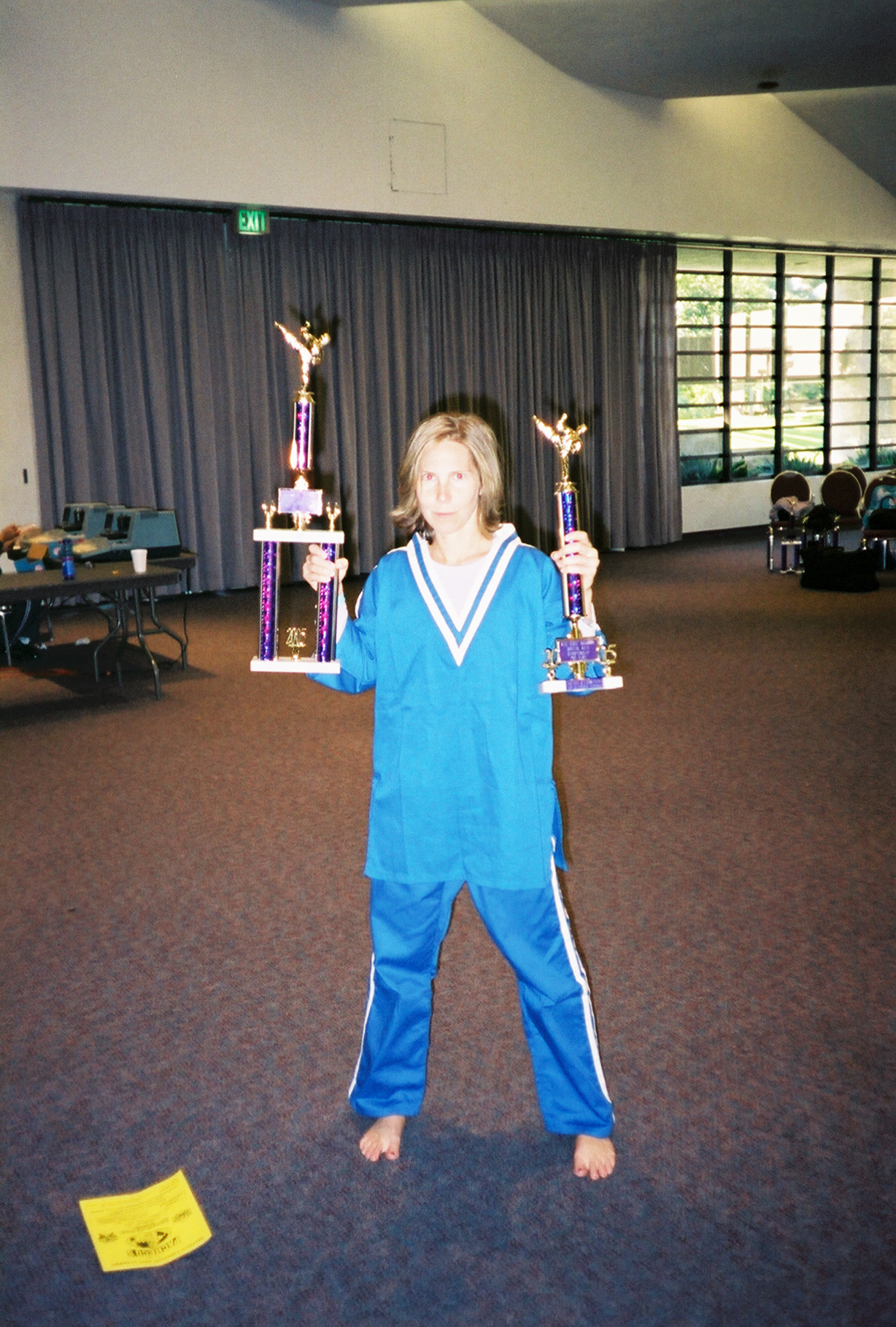 Child in blue sports uniform holding two trophies, standing in a large indoor space with curtains and windows.