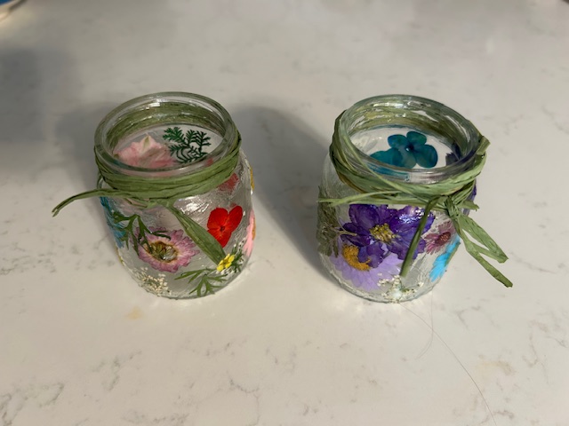 Two small glass jars with painted flowers and tied with green string, viewed from above on a white surface.