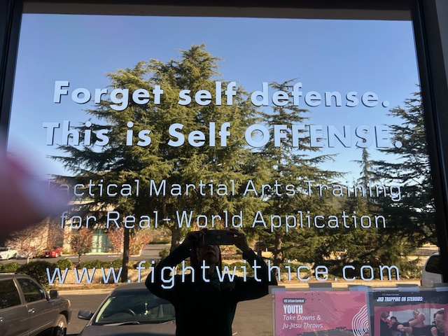 Text on glass window promoting self-defense and martial arts training, with trees and cars outside visible in the background.