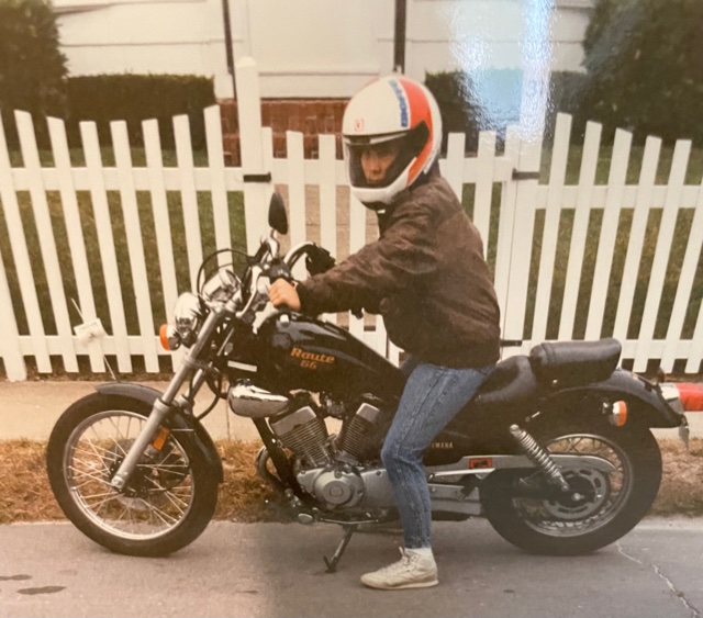 Person wearing a helmet sitting on a black motorcycle on a street, with a white picket fence in the background.