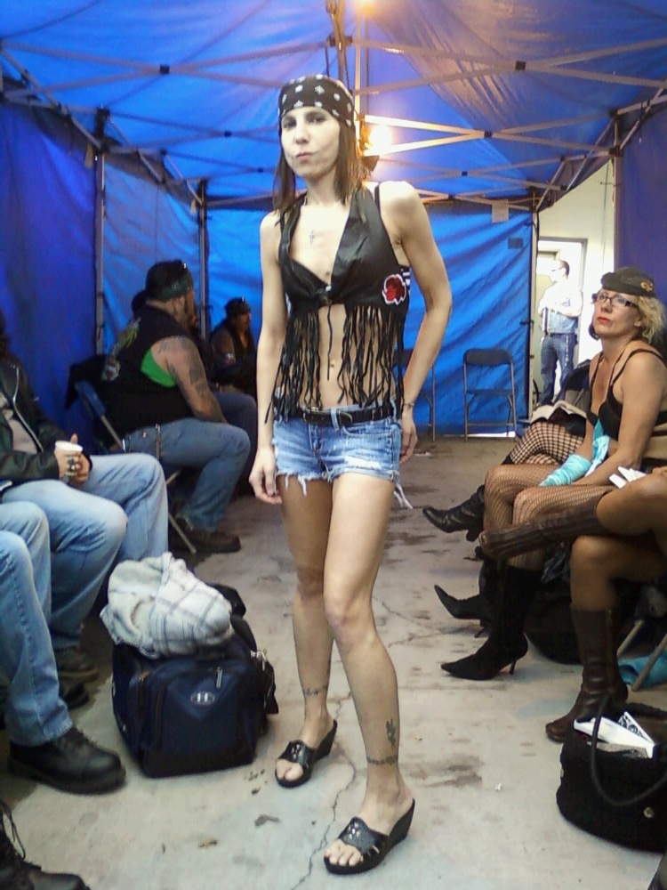 Young woman in a black fringed top, denim shorts, and sandals walking in a tented area with seated people around.