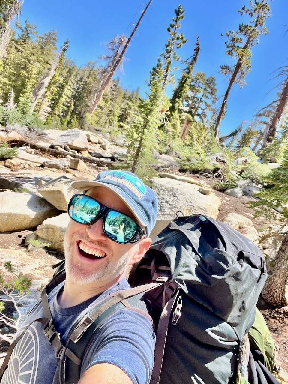 Smiling man wearing sunglasses and a cap, carrying a backpack outdoors in a forested area with rocks and trees.