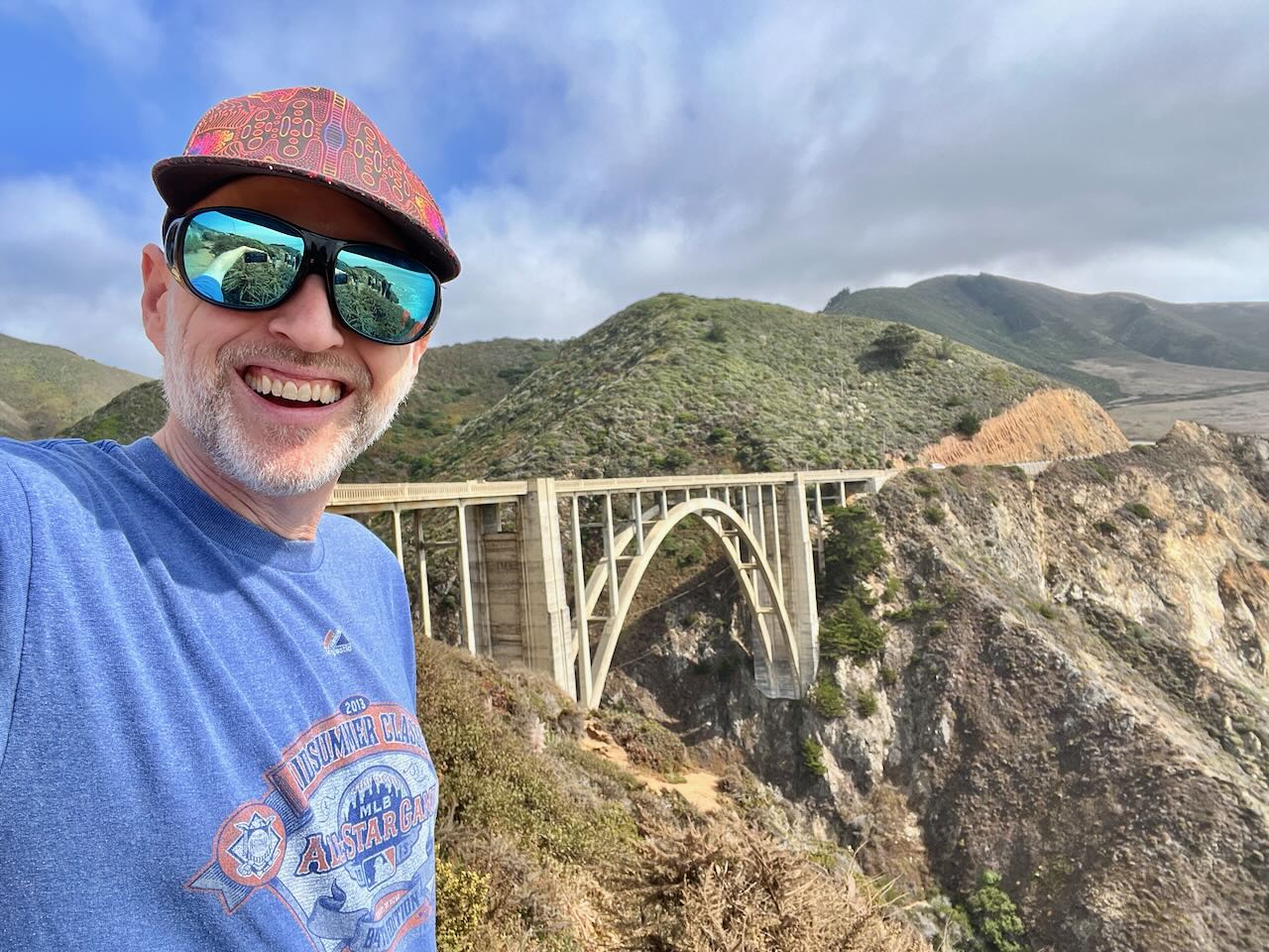 Smiling man wearing sunglasses and a hat takes selfie with a bridge and green hills in background.