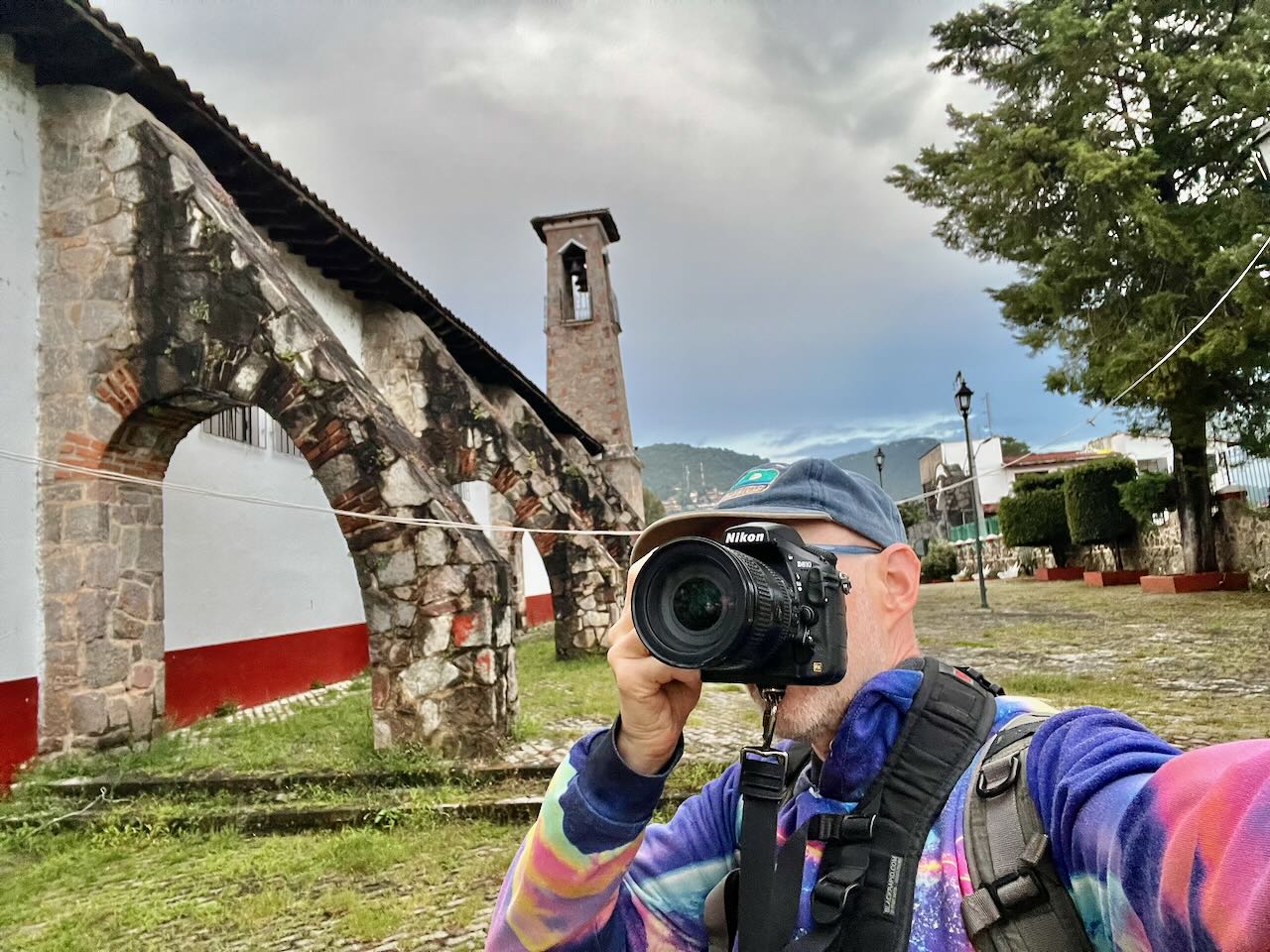 Person taking a photo outdoors with a camera, wearing a cap and colorful jacket, in front of an old stone structure and trees.