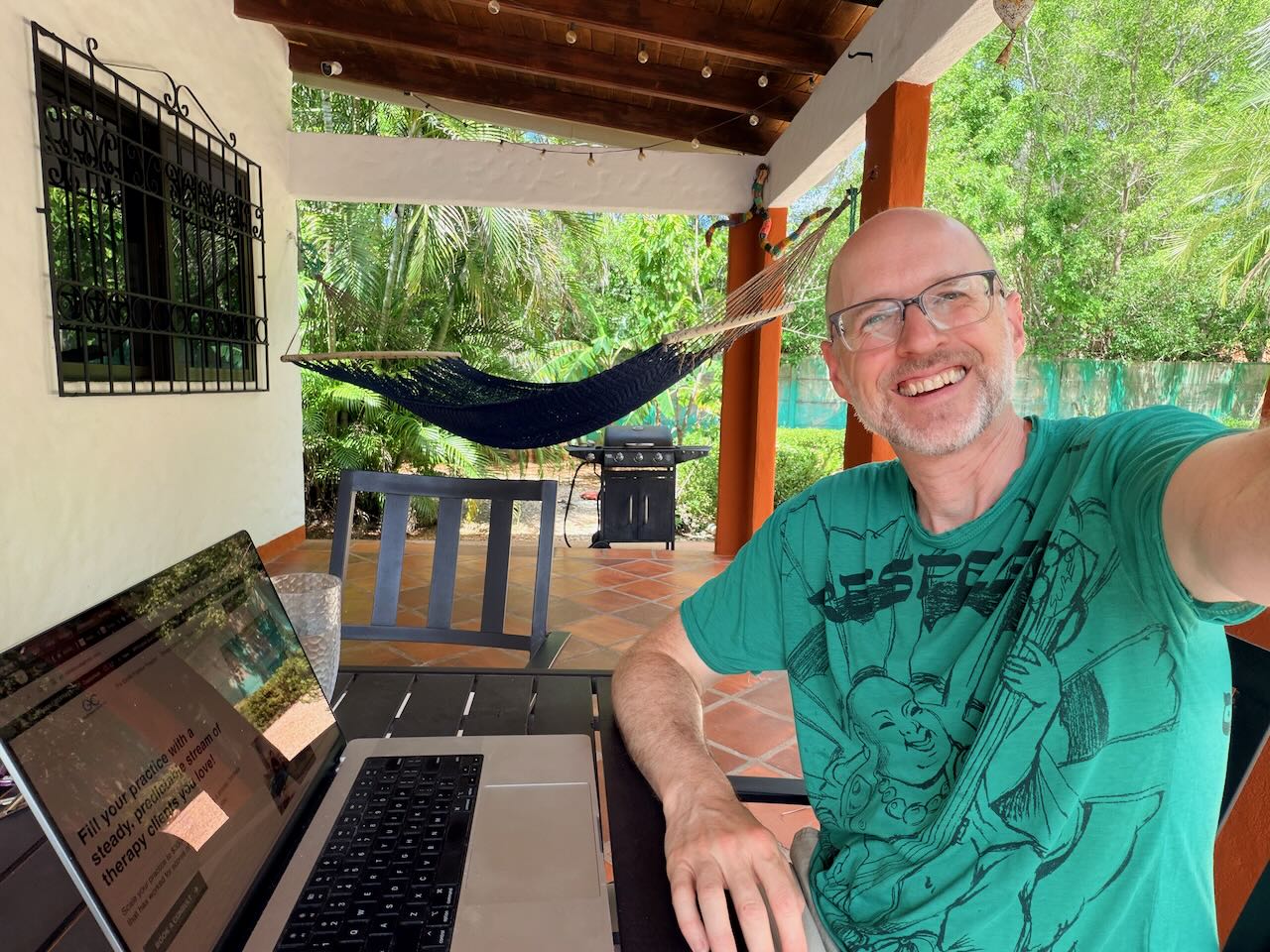 Smiling man with glasses in a green T-shirt sitting outdoors with a laptop, hammock, and trees in background.
