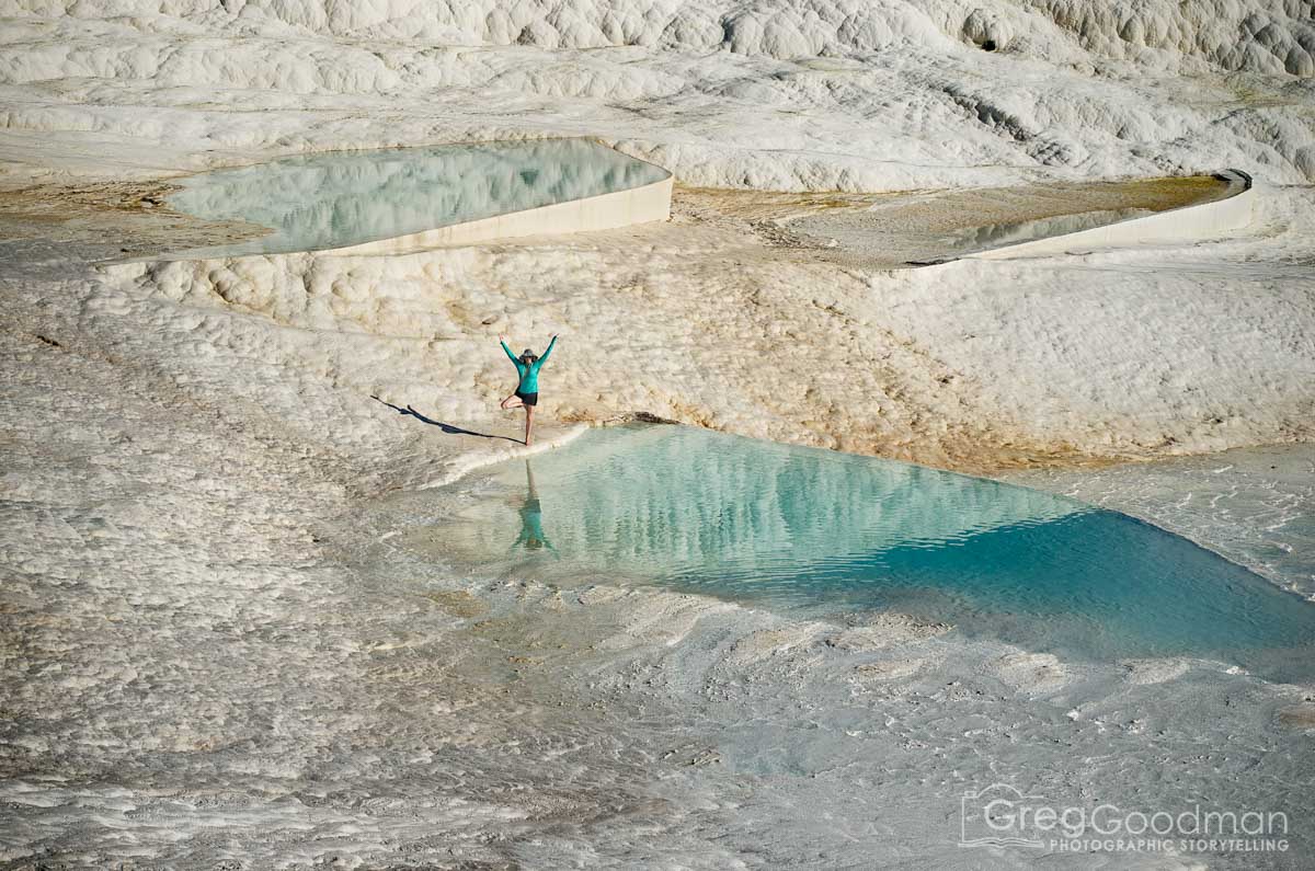 Person standing with arms raised on rocky terrain near turquoise pools of water, landscape appears arid and barren.