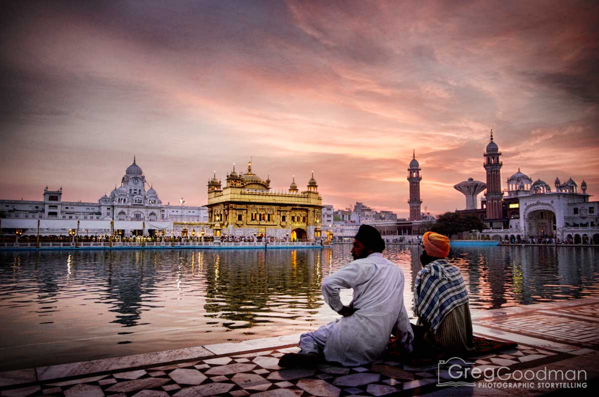 Two people sit by a river at sunset, with a golden temple and other buildings reflected in the water.