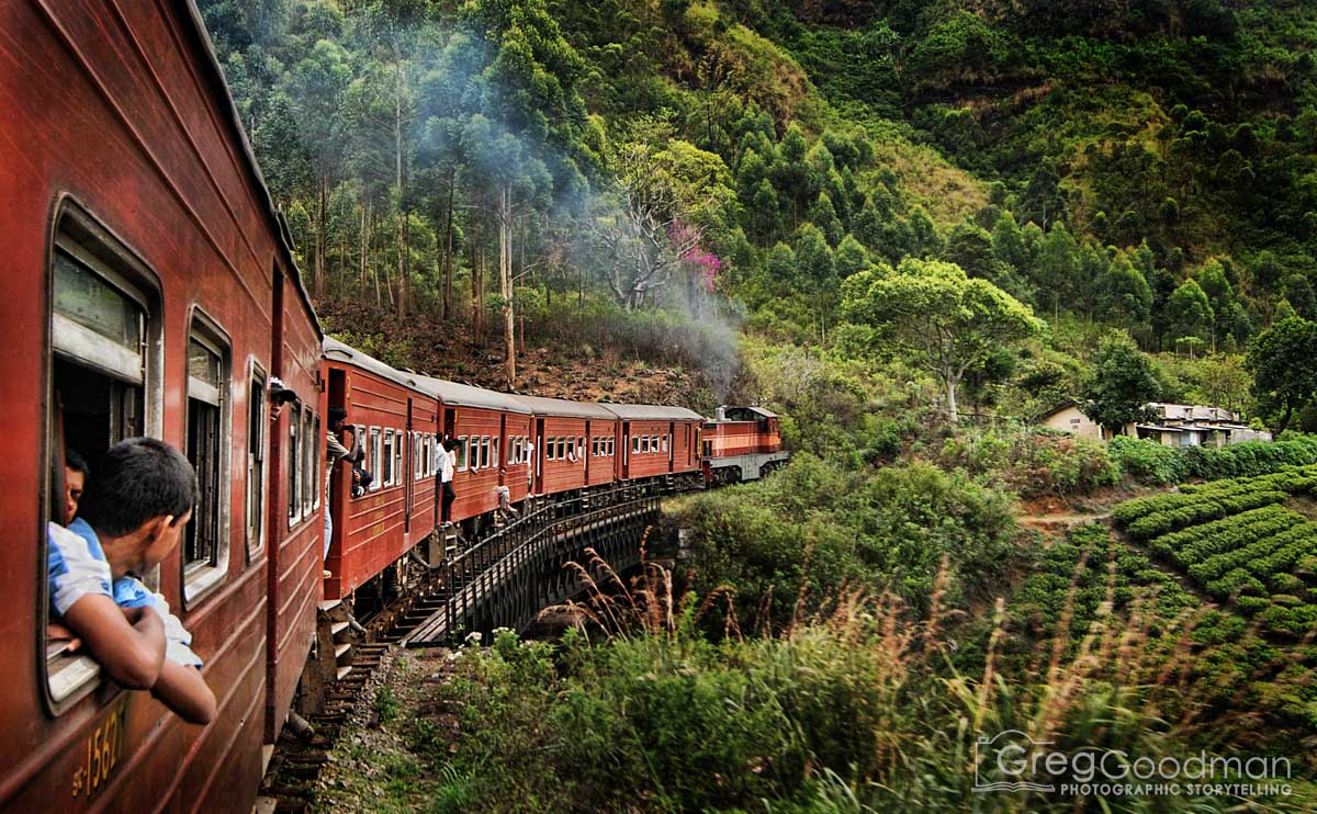 Red train with children looking out windows travels through green hilly landscape with trees and small buildings.