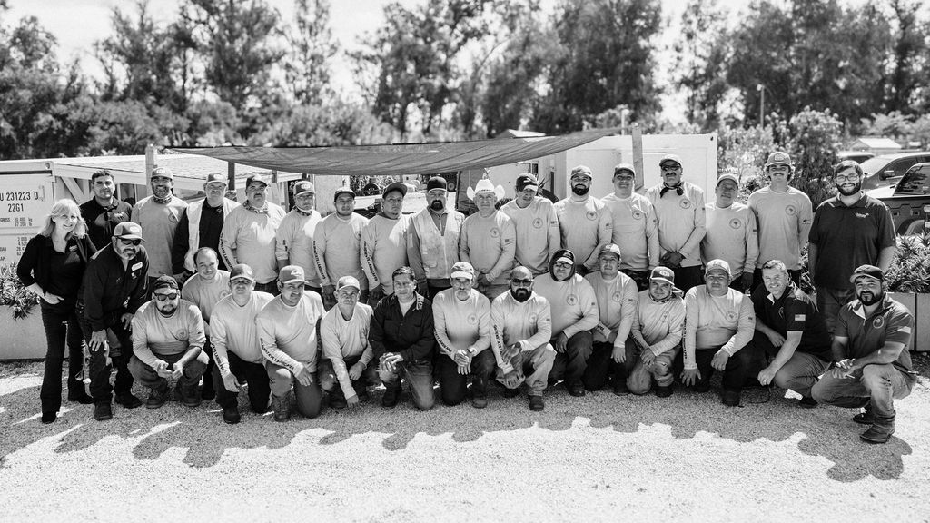 Group of people outdoors, some standing and some kneeling, with trees and a building in the background.