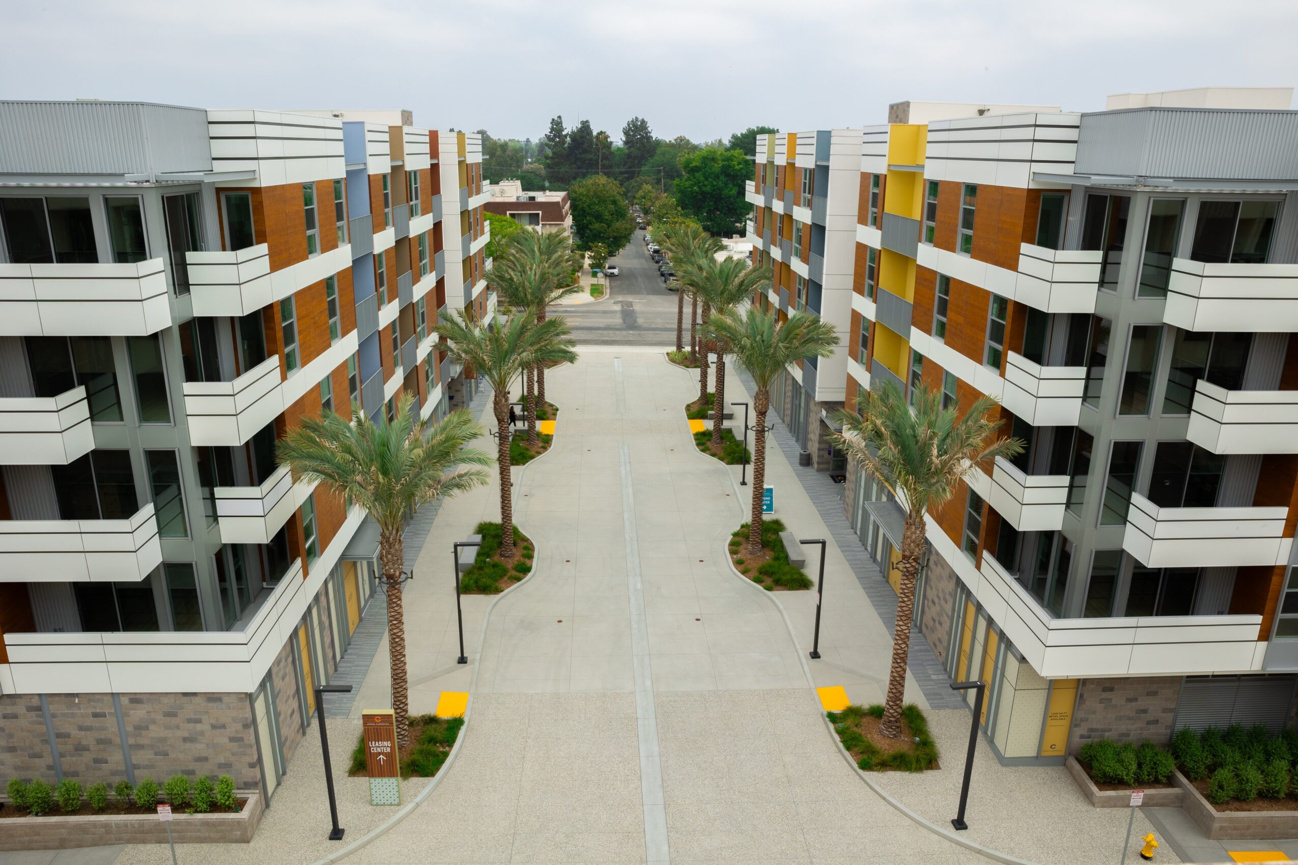 View of a modern residential complex with palm trees lining a paved walkway between multi-story buildings, under a cloudy sky.