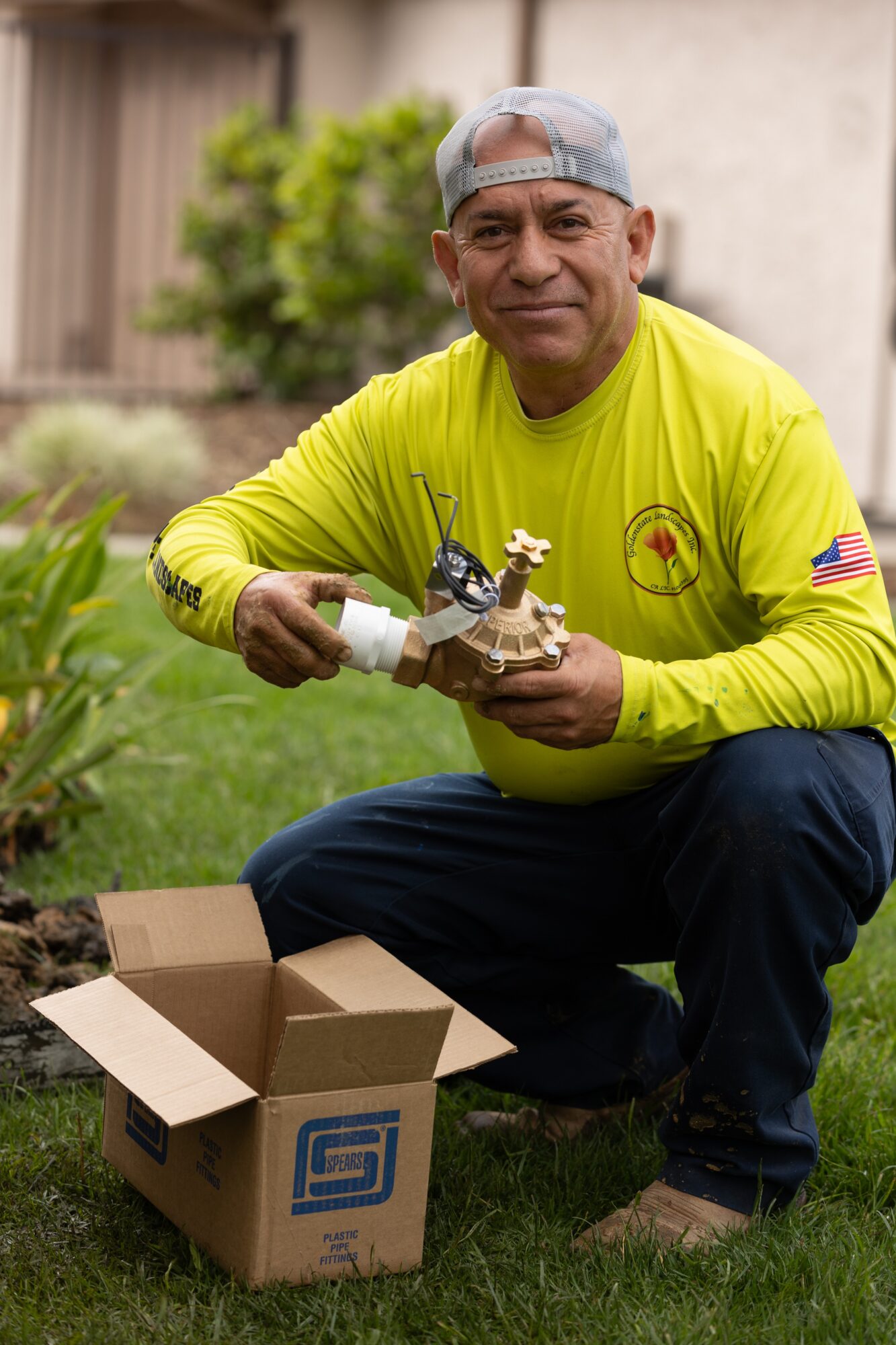 Man in yellow shirt and gray cap planting a small tree outdoors, with an open box nearby.