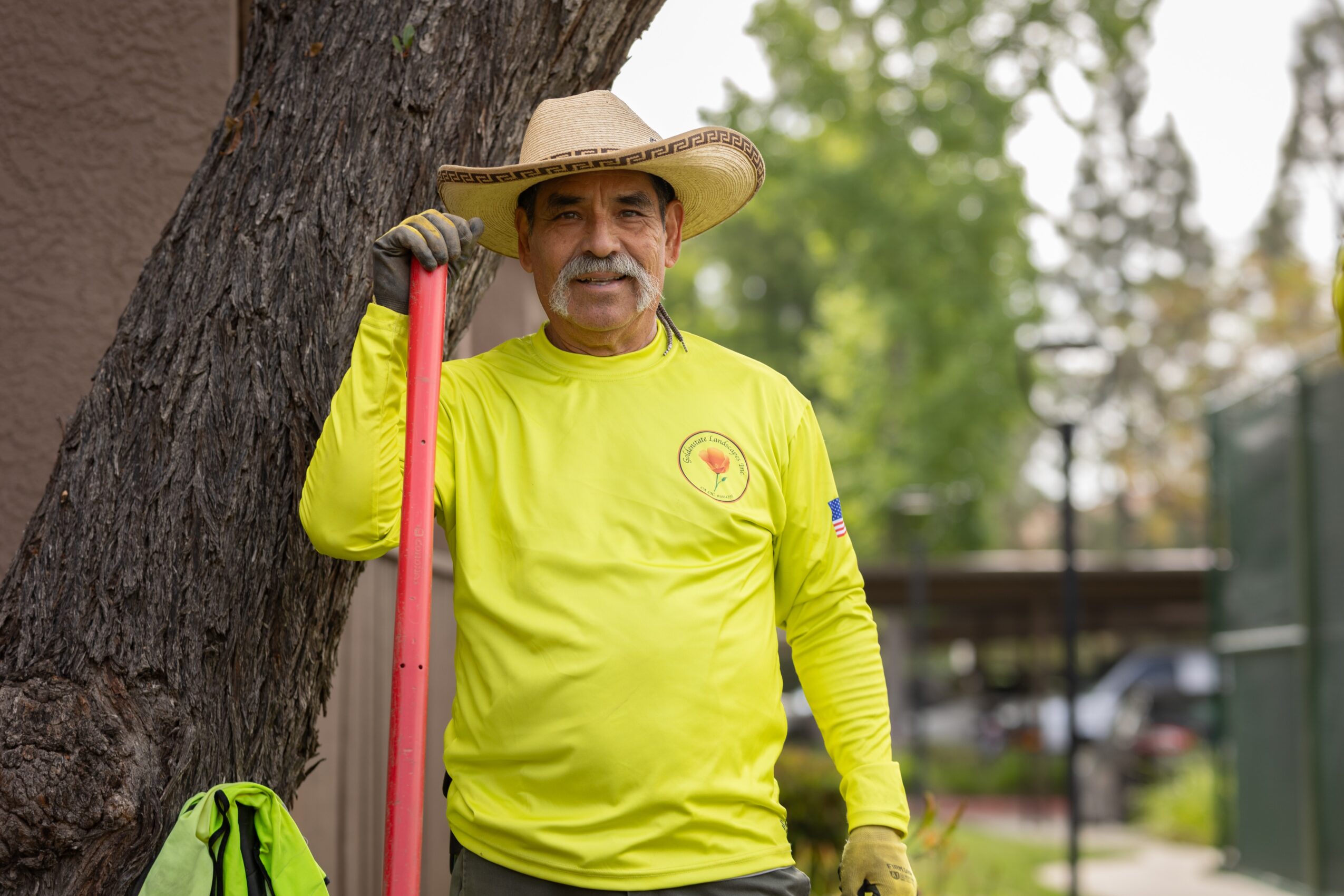 Older man in bright yellow shirt and straw hat leaning against a tree outdoors, holding a pink gardening tool, with greenery in background.