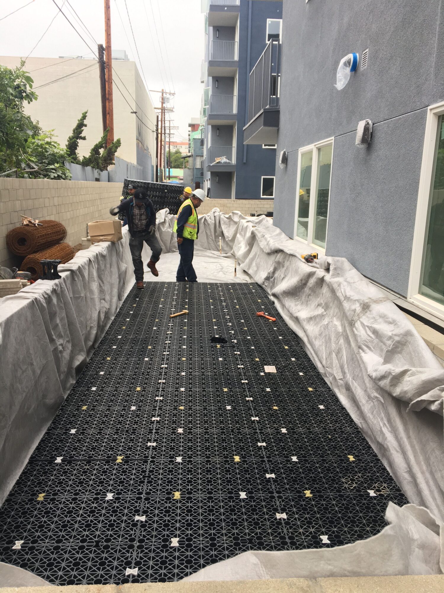 Construction worker in safety gear walking along a black patterned carpet on a building site, with gray walls and windows nearby.