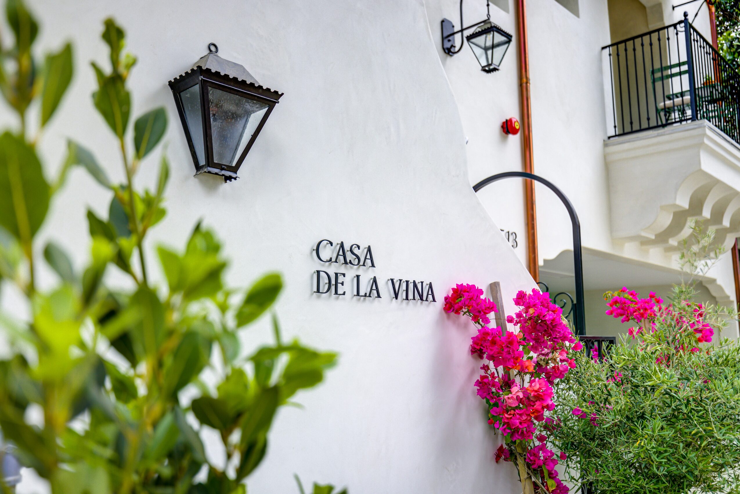 White building with black lantern, pink flowers, and a balcony, with a sign reading 'Casa de la Vina'.