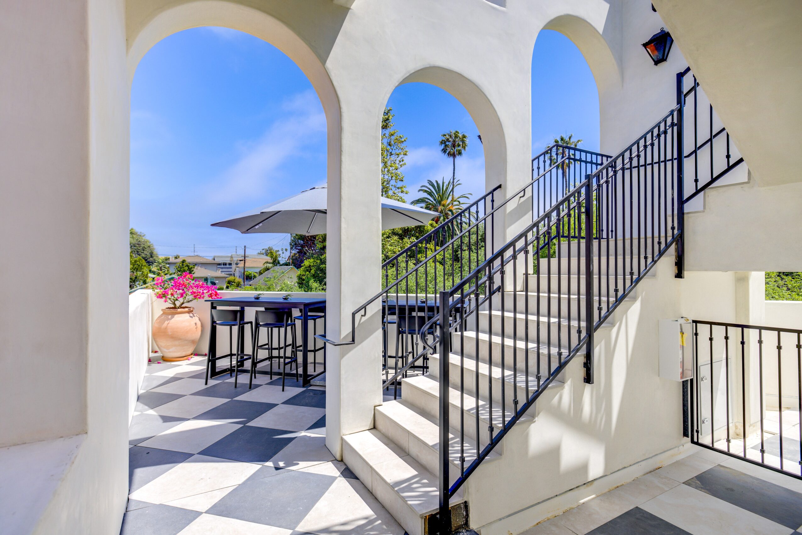 Outdoor patio with checkered floor, table with umbrella, stairs, and arches, overlooking trees and a blue sky.