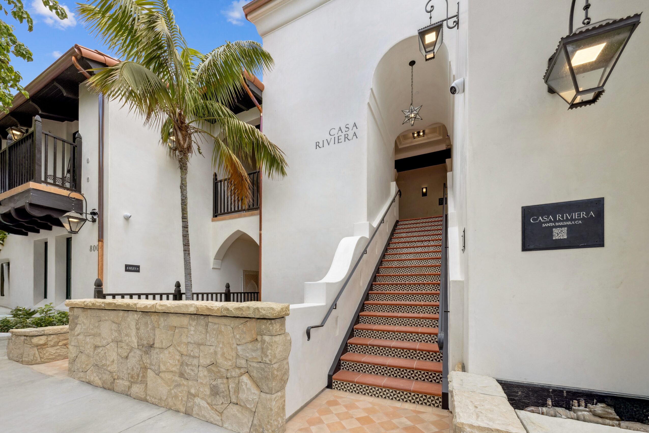 White building with stairs leading to a door, palm trees, and a stone wall in front, sunny sky above.