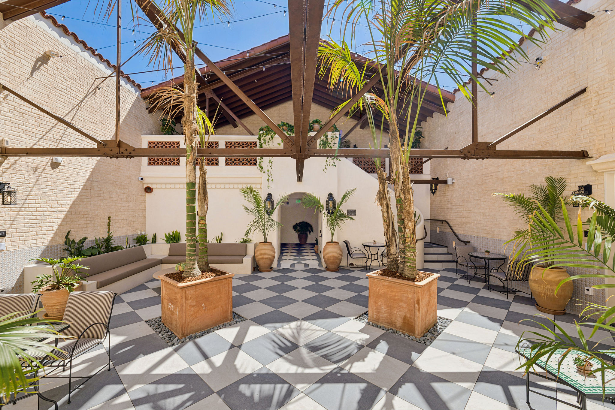 Outdoor patio with checkered floor, potted plants, and palm trees, enclosed by white walls and a wooden roof structure.
