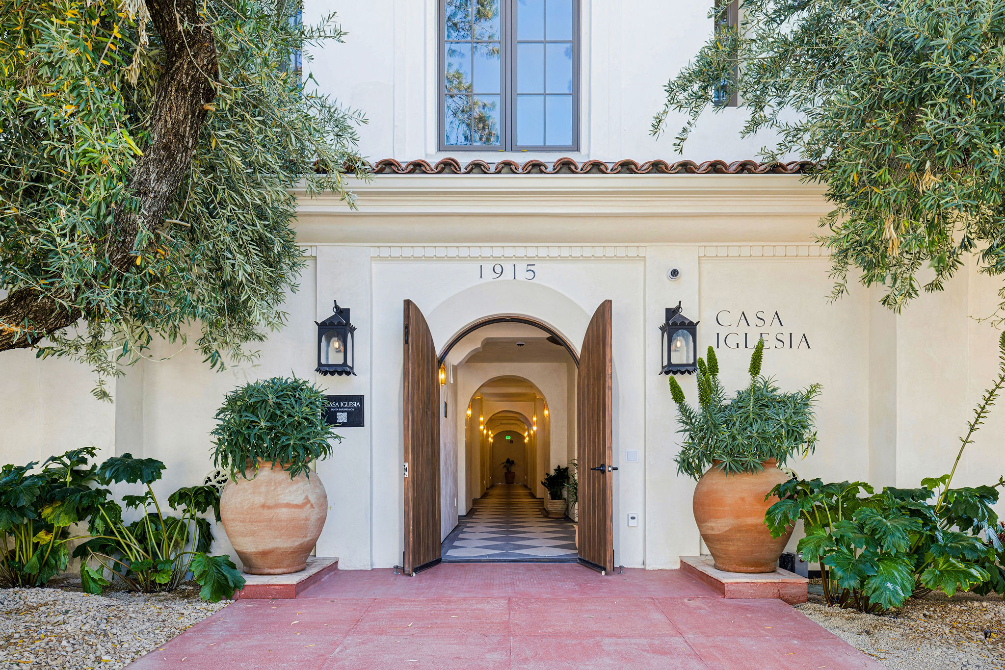 Entrance to a building with an arched doorway, potted plants on either side, and a white wall with a sign reading 'Casa Iglesia'.