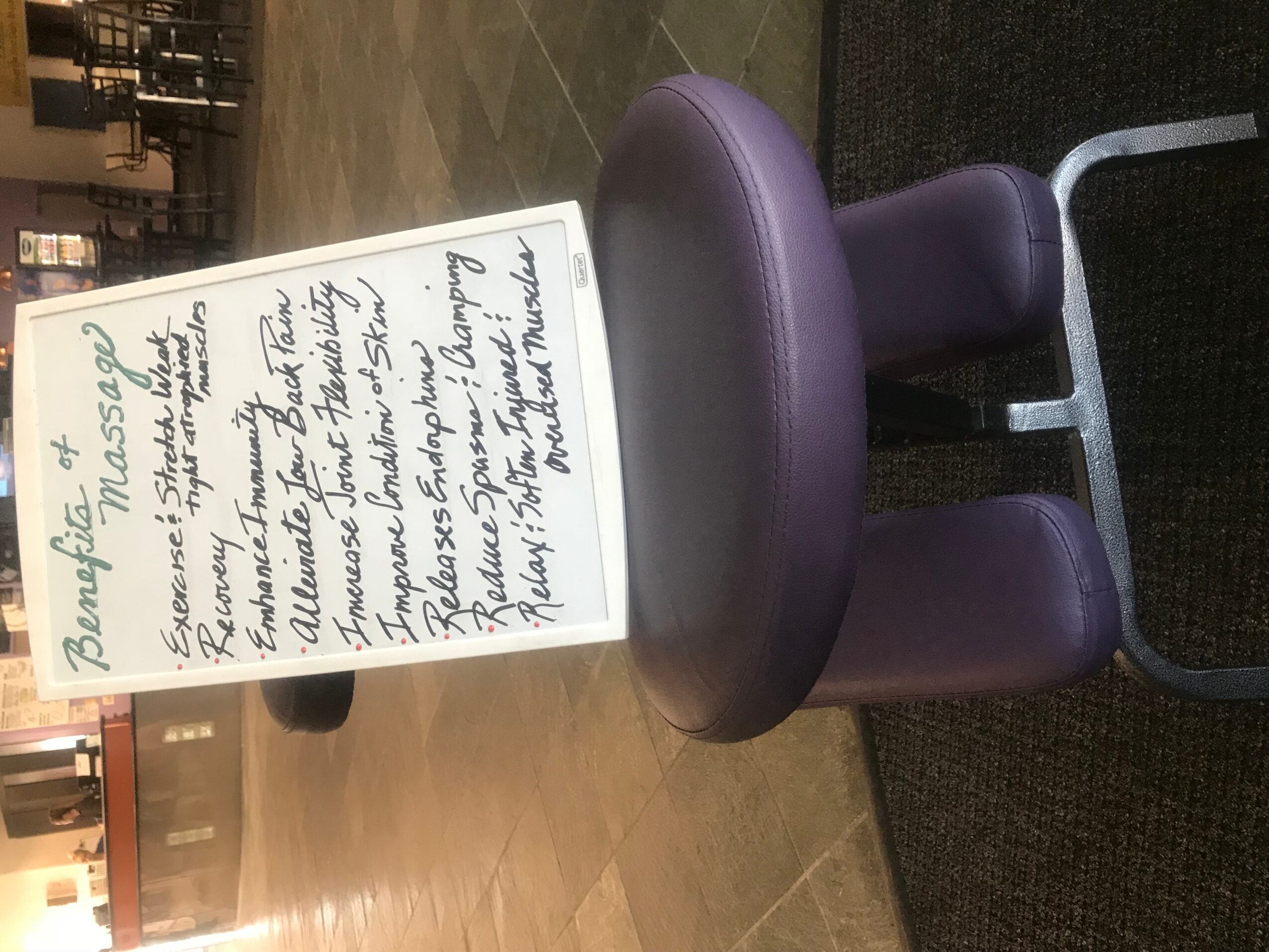 A purple massage chair with a whiteboard sign in front of it, on a wooden floor in a room with shelves.