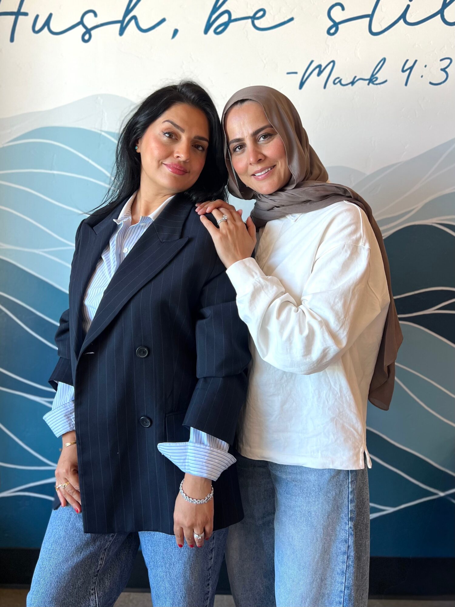 Two women standing close together, smiling, with a blue and white abstract background, one in a dark blazer and the other in a white jacket.