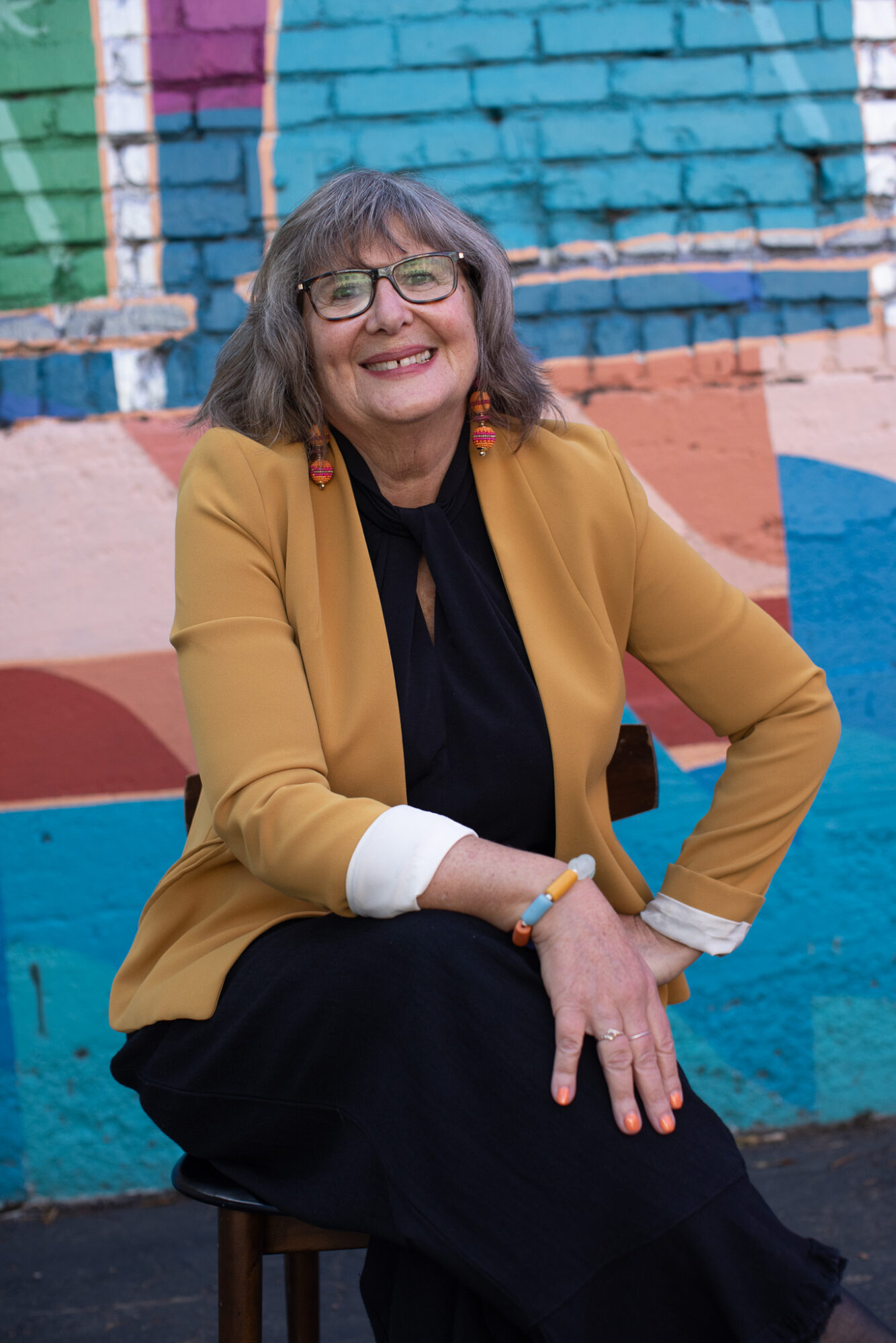 Woman with glasses and shoulder-length hair smiling, sitting on a stool in front of a colorful brick wall, wearing a mustard blazer and black dress.