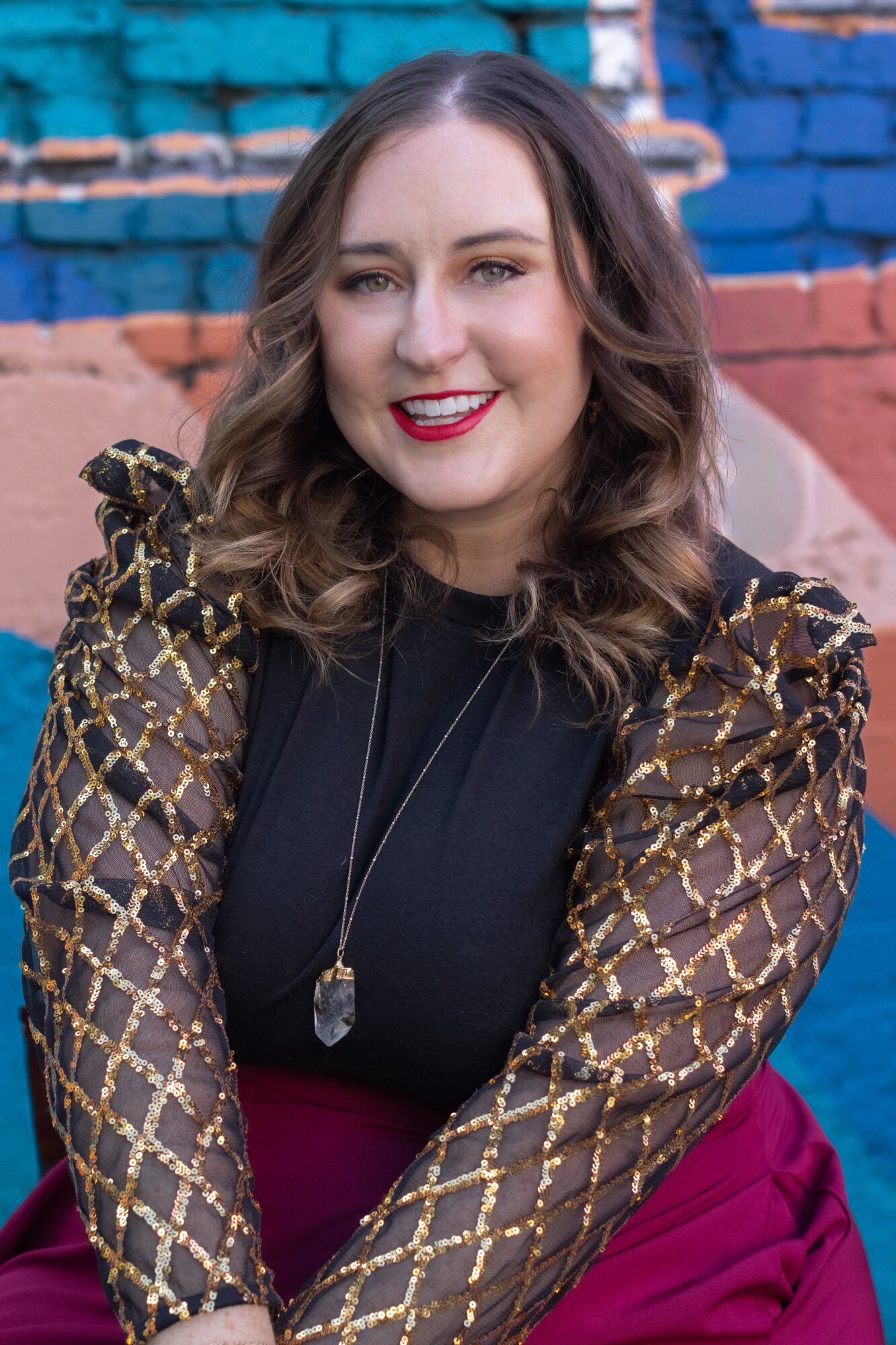 Woman with wavy hair smiling, wearing a black top and patterned jacket, in front of a colorful brick wall.