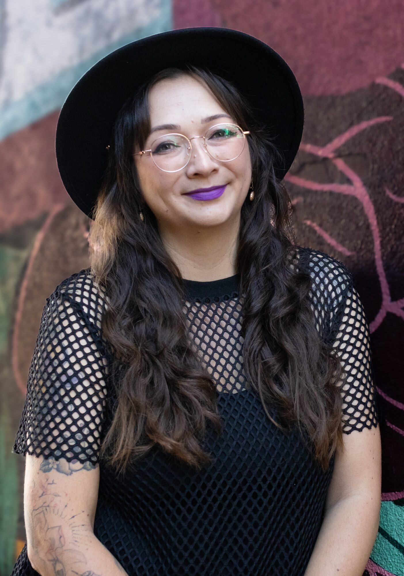 Young woman with long wavy hair, glasses, purple lipstick, wearing a black hat and mesh top, standing outdoors.