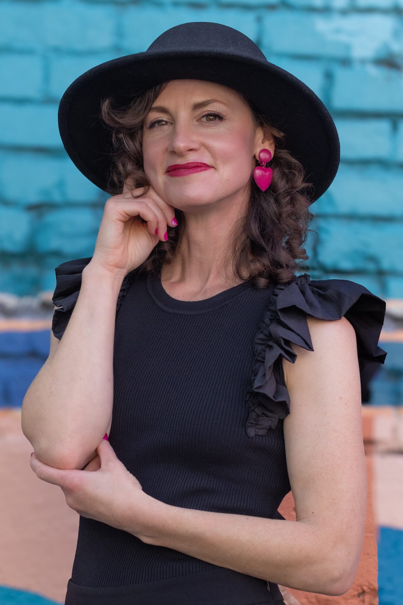 Woman wearing a black hat and dress with ruffled sleeves, pink earrings, standing against a blue brick wall.