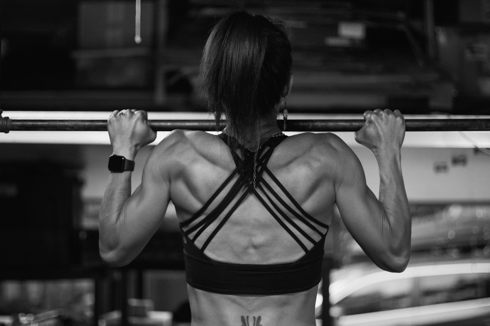 Back view of a woman lifting a barbell in a gym, showing muscular back and shoulder muscles, black sports bra, and workout setting.