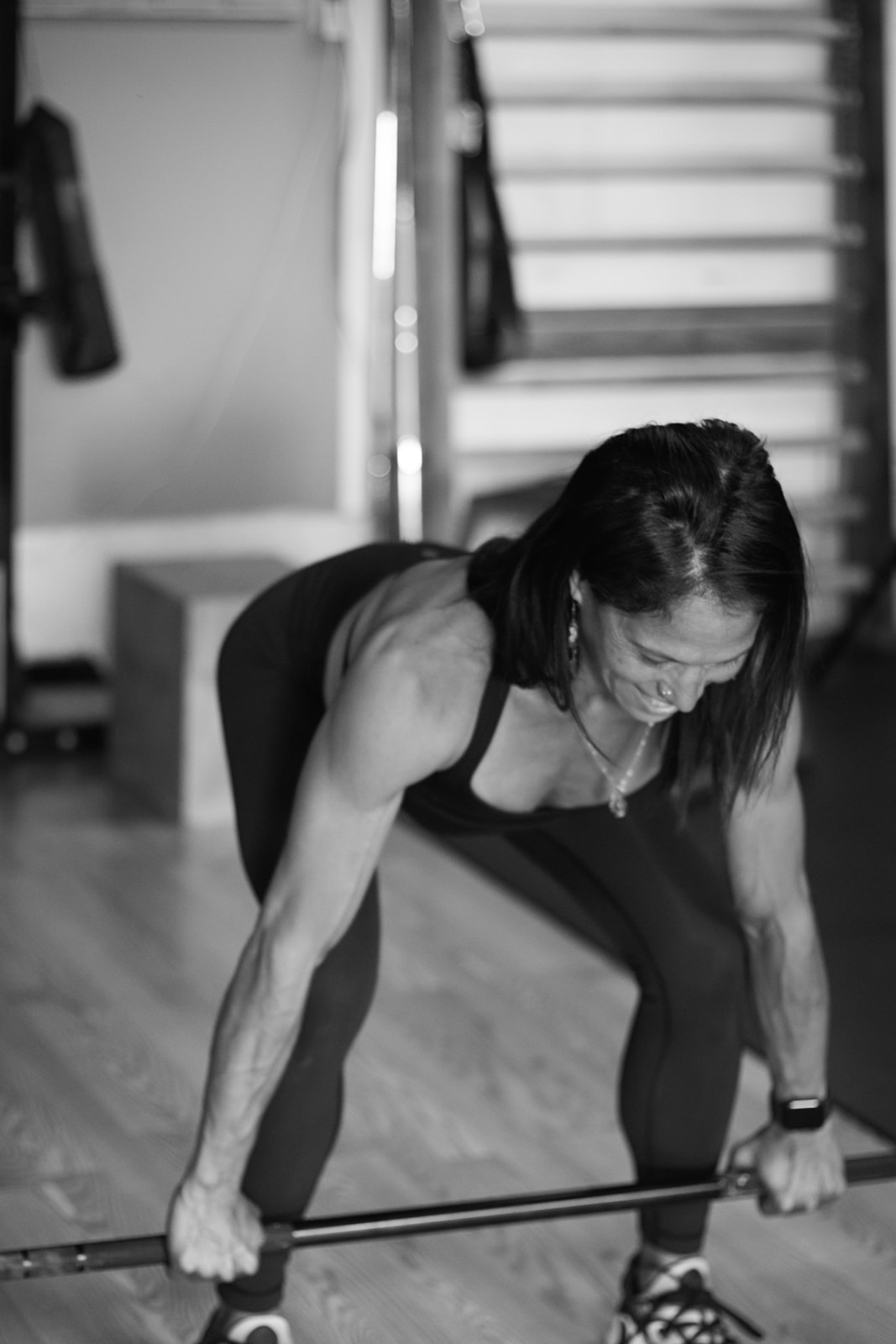 Woman in workout clothes bending forward holding a barbell in a gym, stairs in background