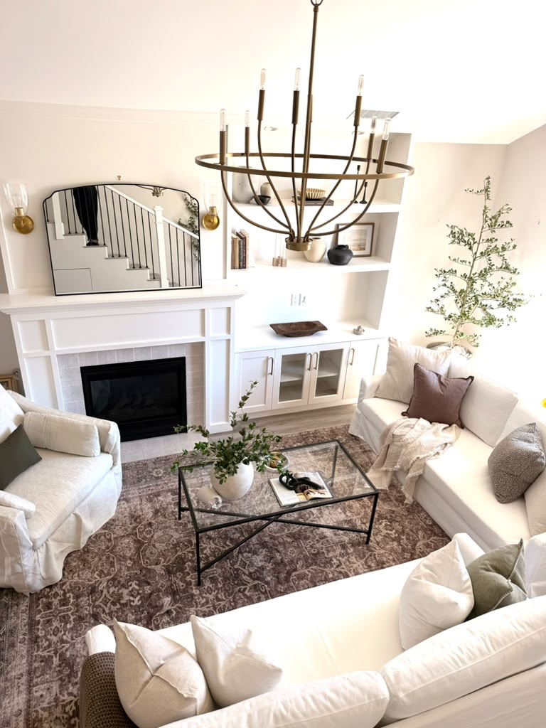 Living room with white sofas, a glass coffee table, a fireplace, and a chandelier, decorated with plants and pillows.