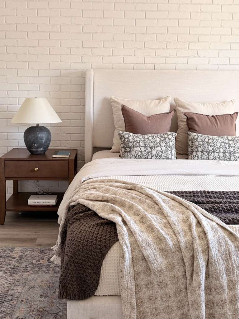 Bed with pillows, beige and brown blanket, white headboard, brick wall, nightstand with lamp, and books.