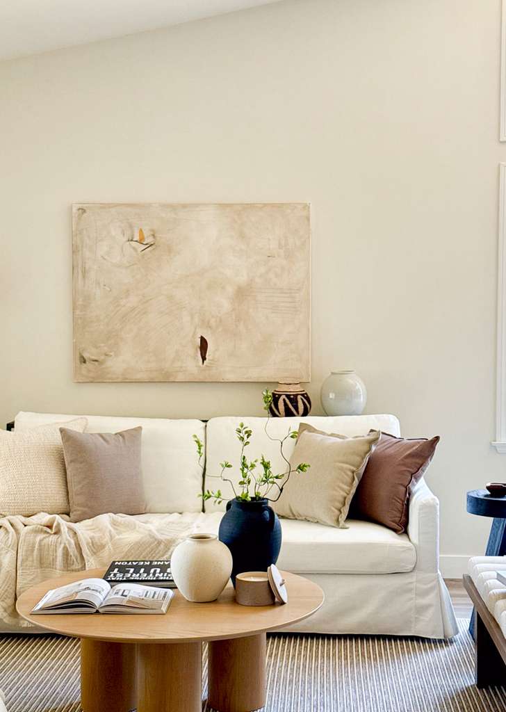 Living room with a white sofa, beige and brown cushions, a wooden coffee table with books and vases, and a large abstract painting on the wall.