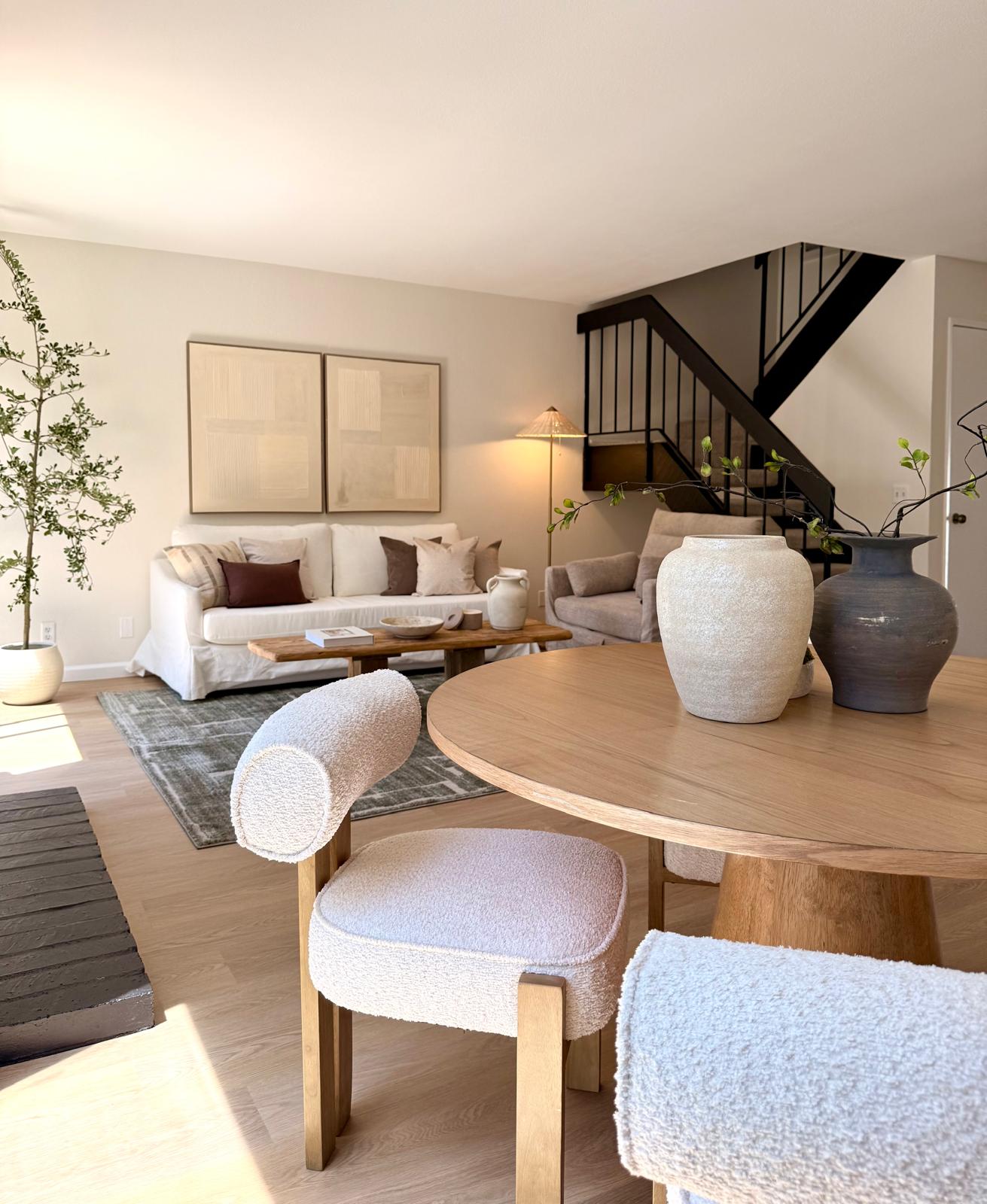 Living room with white sofa, armchair, round wooden table with vases, staircase, and potted plant, bright and modern decor.