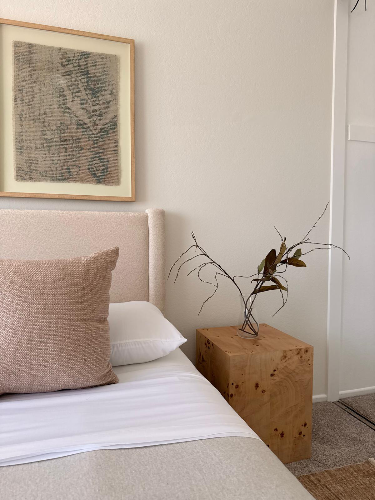 Bedroom corner with bed, pillow, framed artwork, and a wooden side table with a branch arrangement.