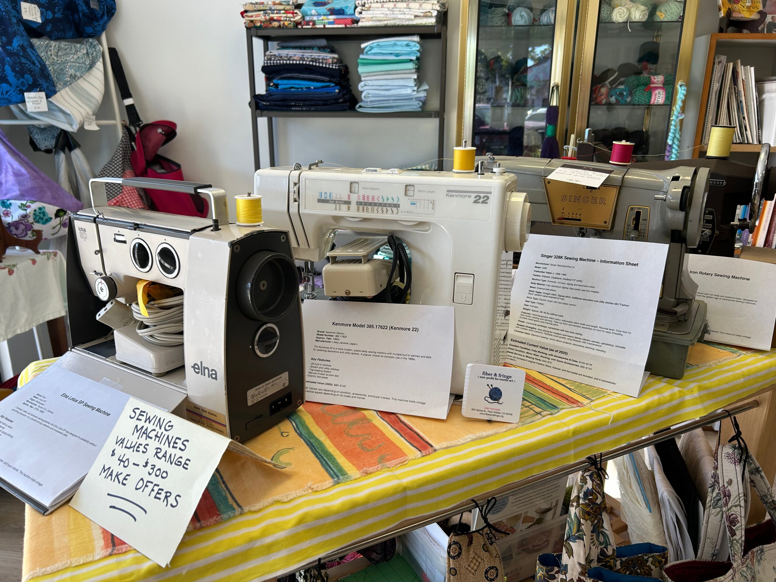 Display of sewing machines, accessories, and informational signs on a table in a room with shelves and cabinets in the background.