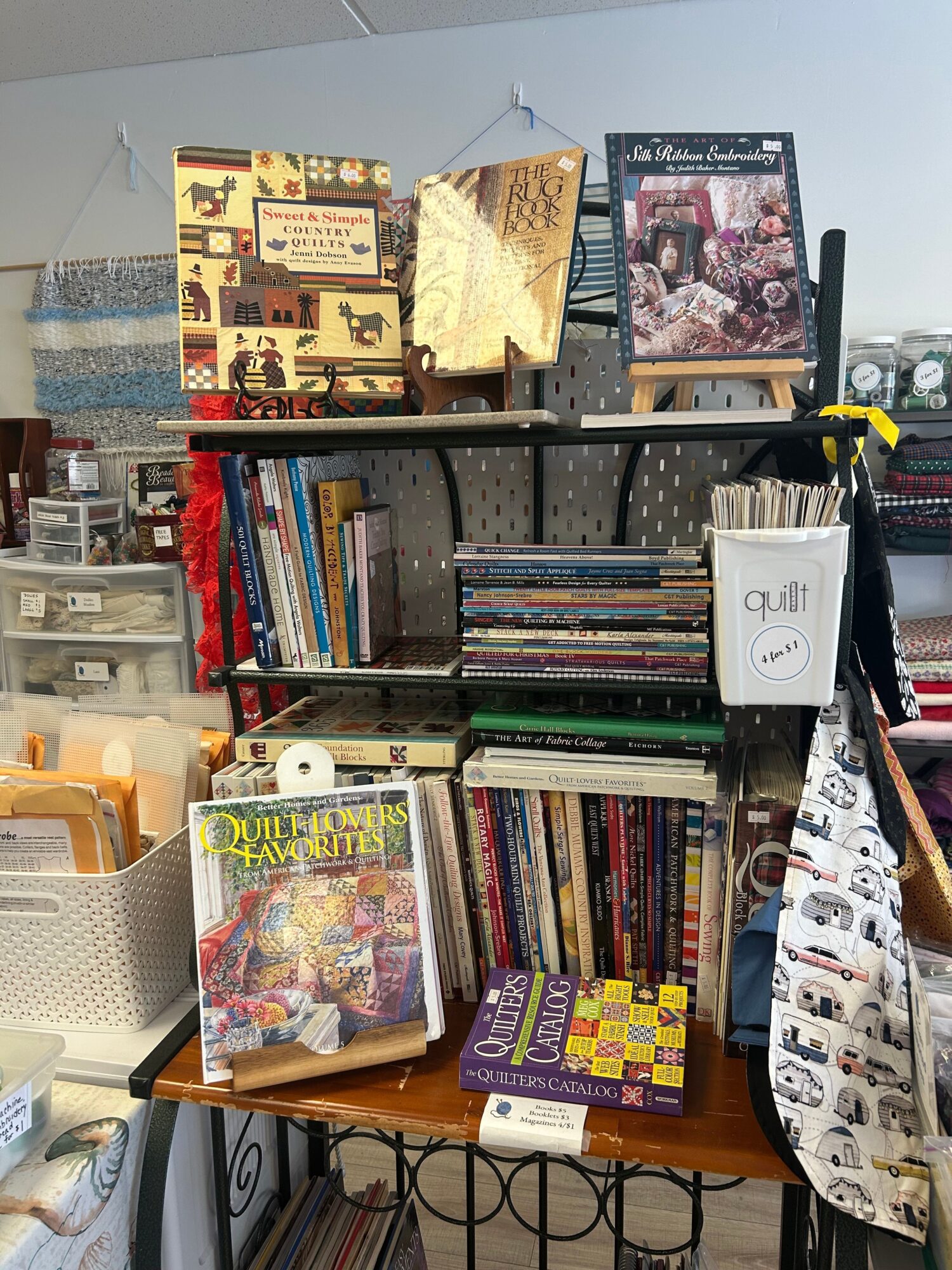 Bookshelves filled with books, magazines, and decorative items, with a framed picture on the wall behind, and a table with more books and a bag.