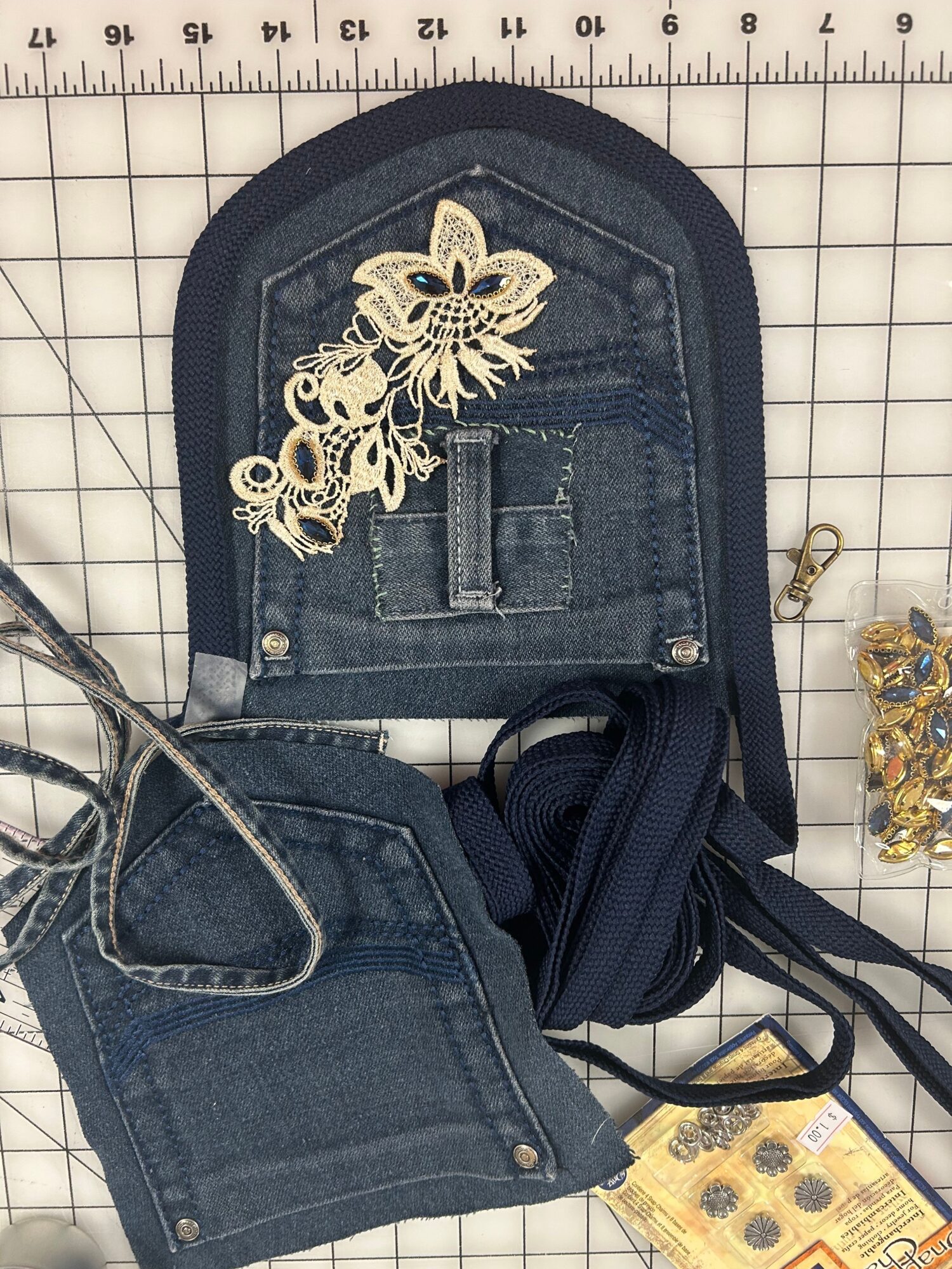 Dark denim bag with lace and floral embroidery, placed on a grid surface with a ruler, and some fabric and accessories nearby.
