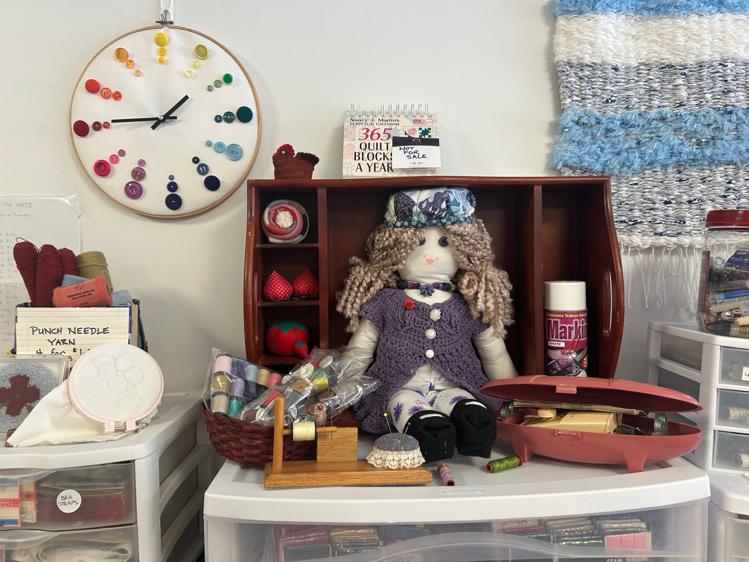 Doll with curly hair and dress sitting on a white table, surrounded by craft supplies and a wall clock, in a craft room.